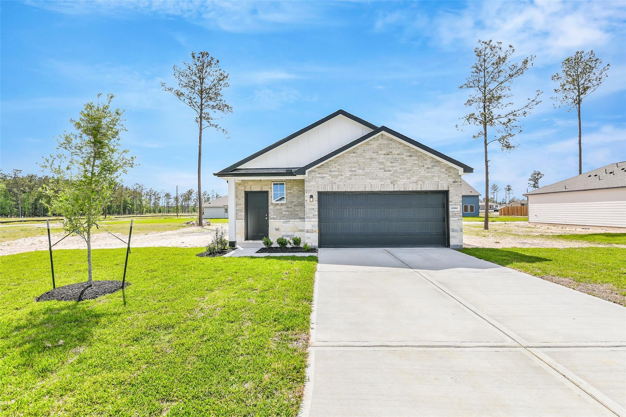 Modern beige brick single-story home with 2-car garage, front porch, lush green lawn, and pine trees in Spring Branch Crossing, Conroe, Texas