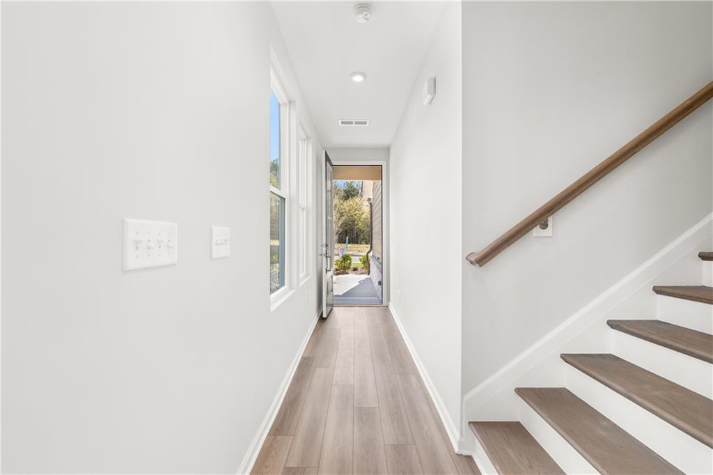 Bright white hallway with light wood floors, oak staircase, large window, and open front door to yard in The Cary A home, Winder, Georgia