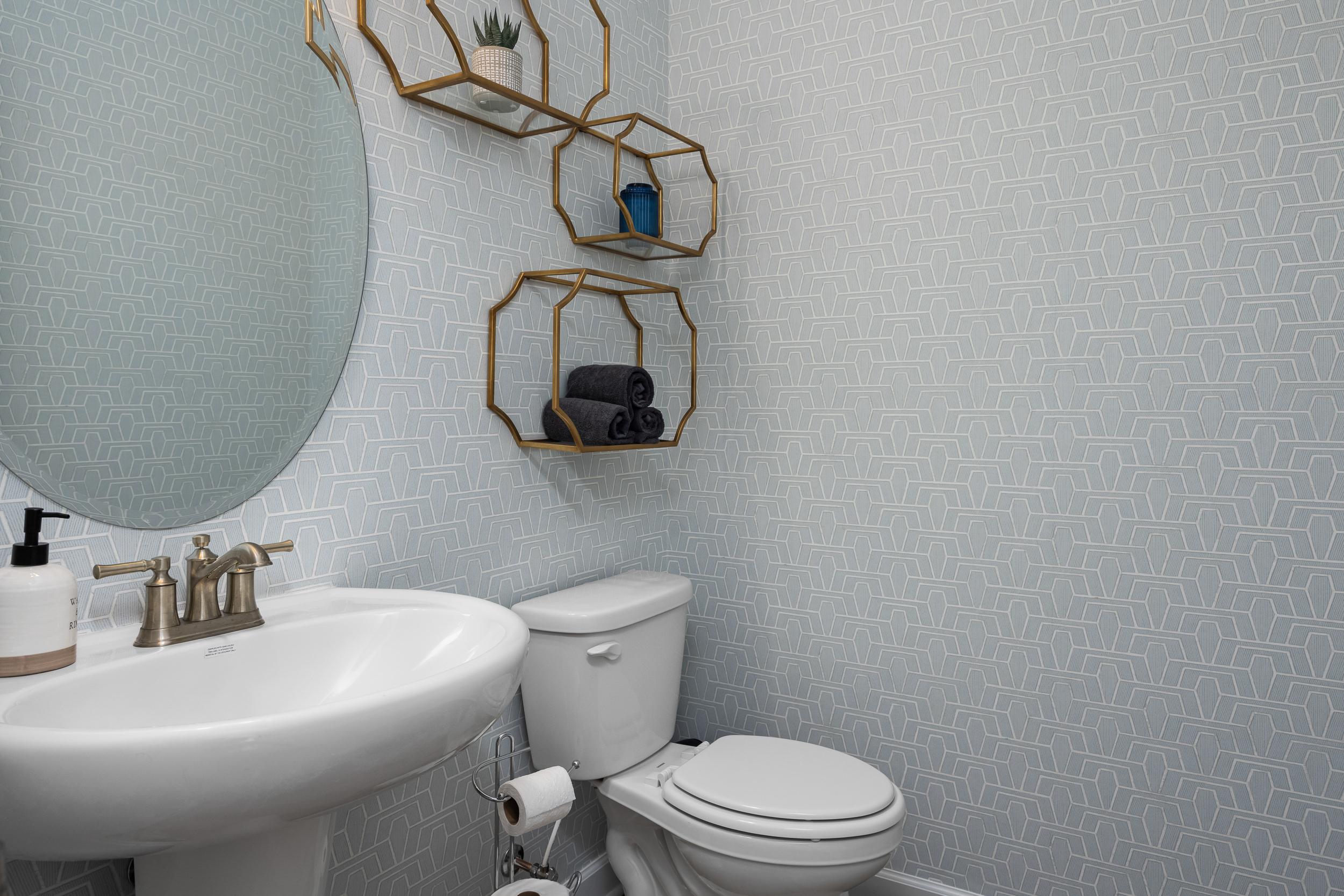 Stylish powder room in The Magnolia A featuring light blue geometric walls, gold geometric shelves with plants and towels, round mirror, white pedestal sink, and toilet