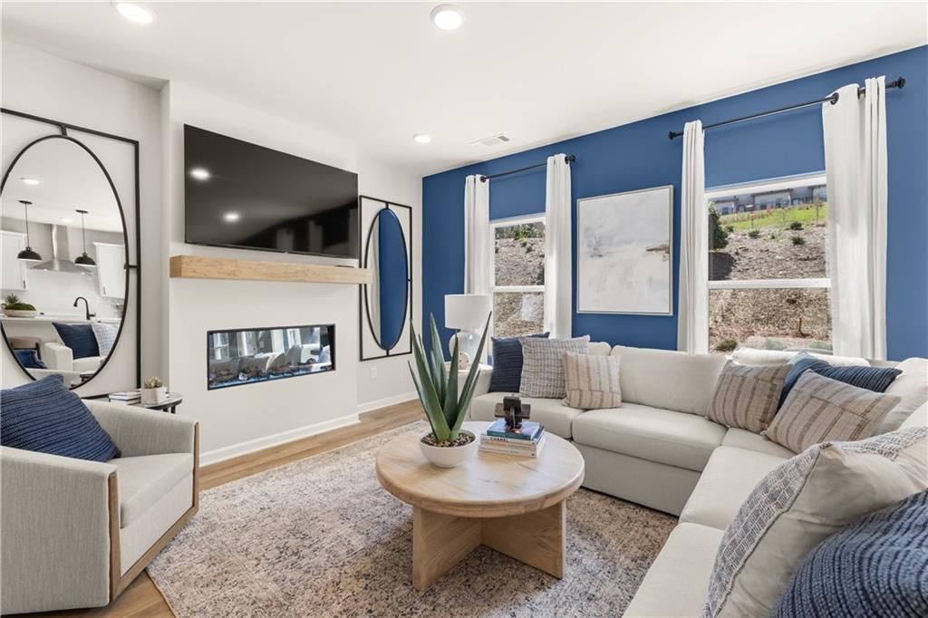 Cozy modern living room with white L-shaped sofa, linear gas fireplace, wall-mounted TV, blue accent wall, and large windows in Davidson Homes The Burton C, Canton, GA