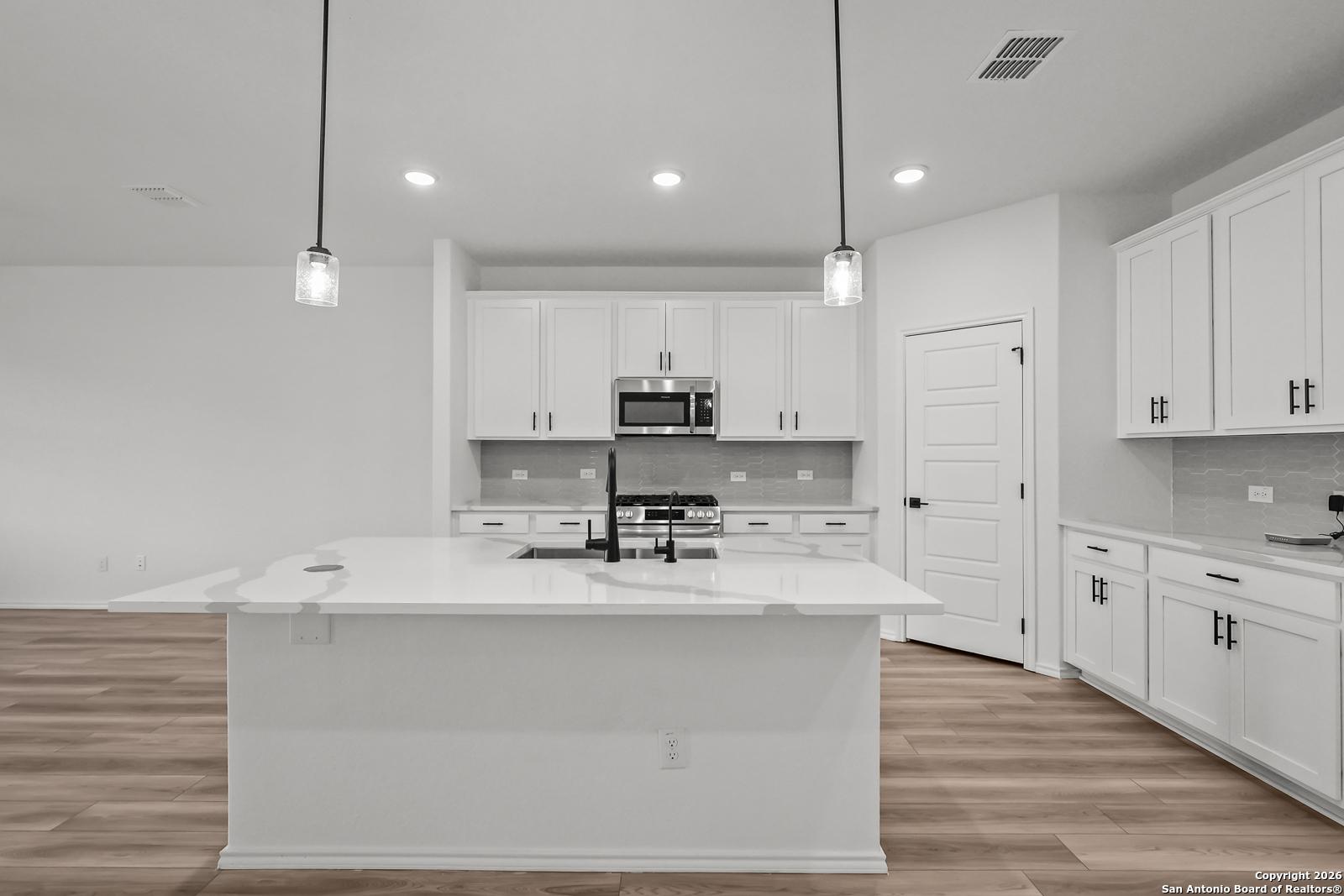 Modern white kitchen with quartz island, farmhouse sink, stainless appliances, and pendant lights in Davidson Homes Douglas E, San Antonio