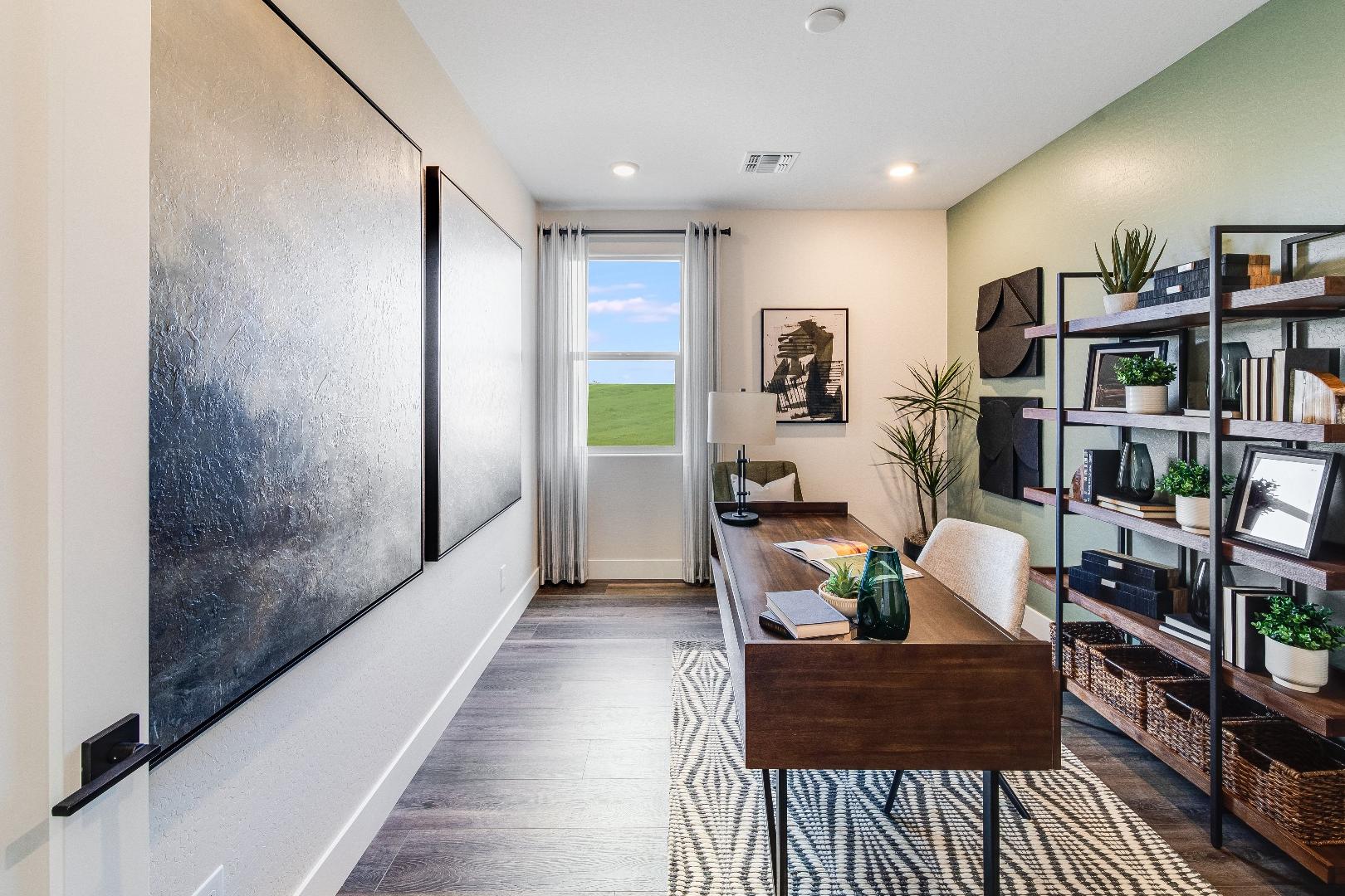 Modern home office in The Boulder II showcasing wooden desk, metal bookshelves, abstract wall art, and sunny window view