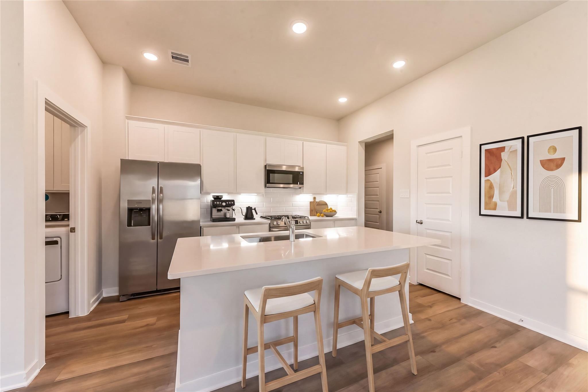 Modern kitchen with white cabinets, stainless steel appliances, quartz island, bar stools, and hardwood floors in Davidson Homes The Costa C, Dayton, Texas