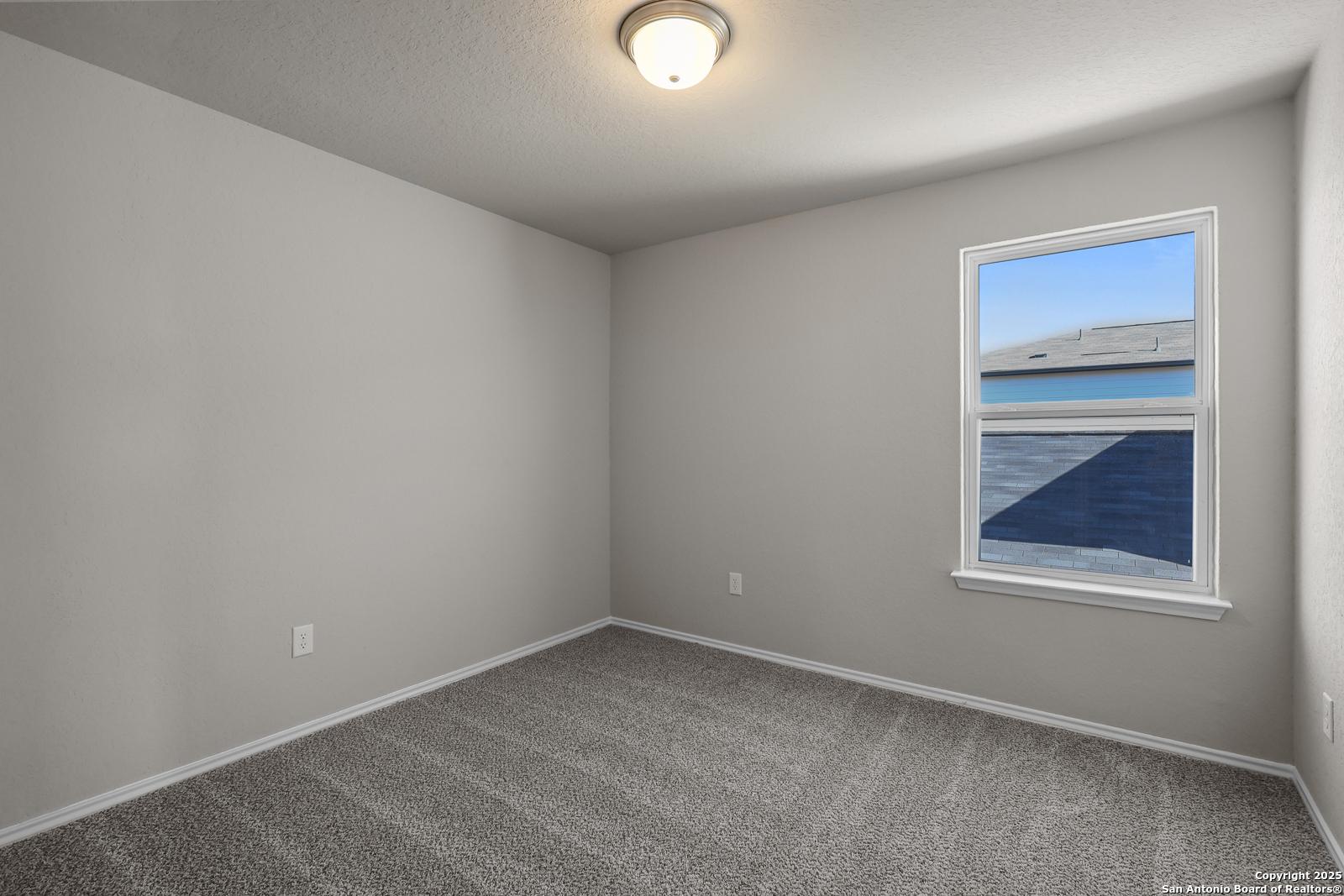 Bright secondary bedroom with light gray walls, plush gray carpet, and large window overlooking blue sky in Davidson Homes The Trinity D, San Antonio