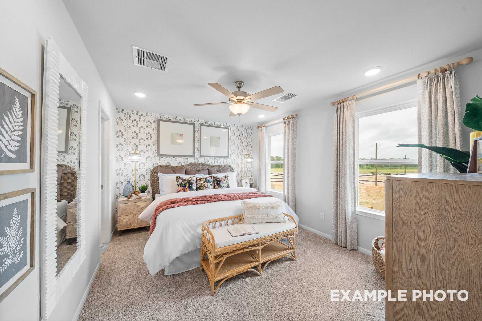 Spacious master bedroom in The Trinity F featuring king bed with red accents, rattan bench, ceiling fan, large window, wooden dresser, and potted ferns