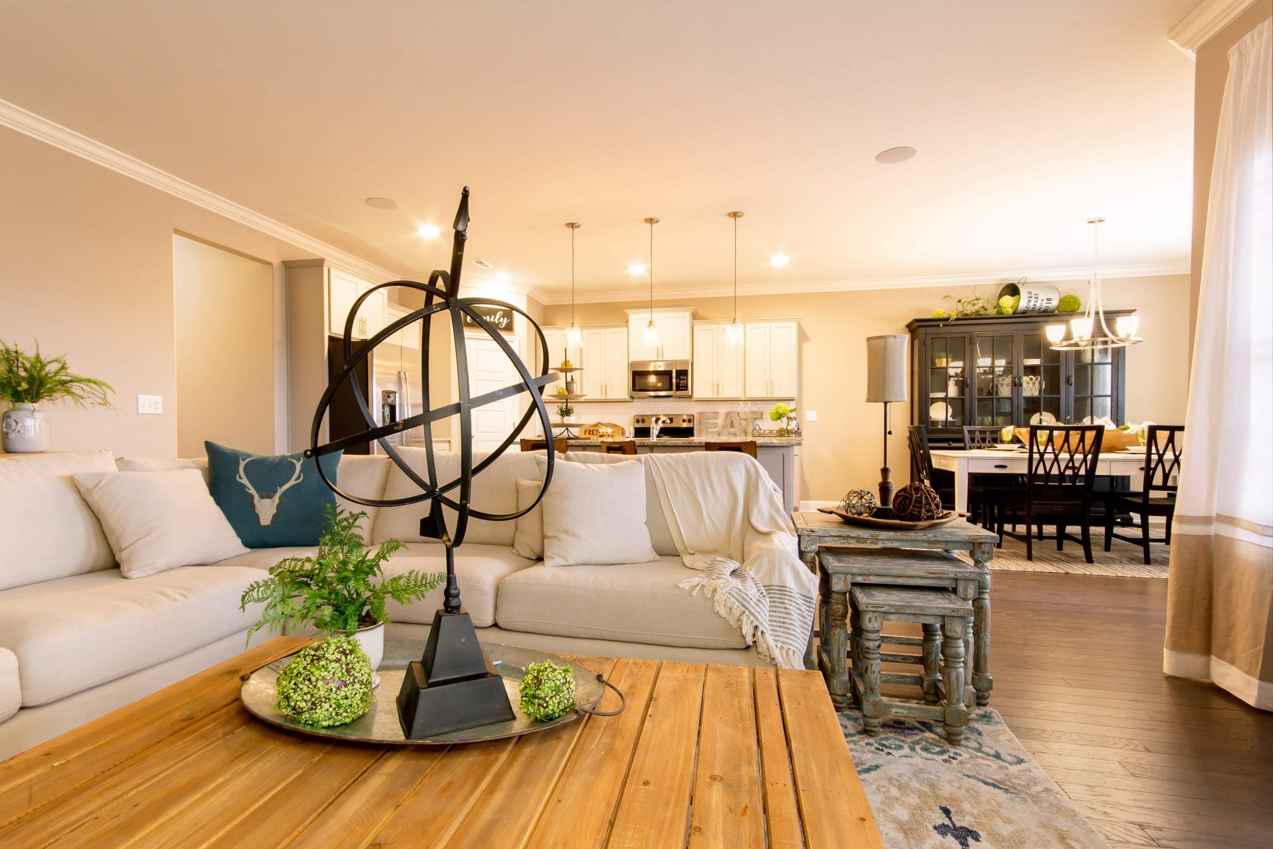Open-concept living room in Bakers Farm home, Priceville AL by Davidson Homes featuring beige sofa, wooden coffee table, and adjacent kitchen with white cabinets