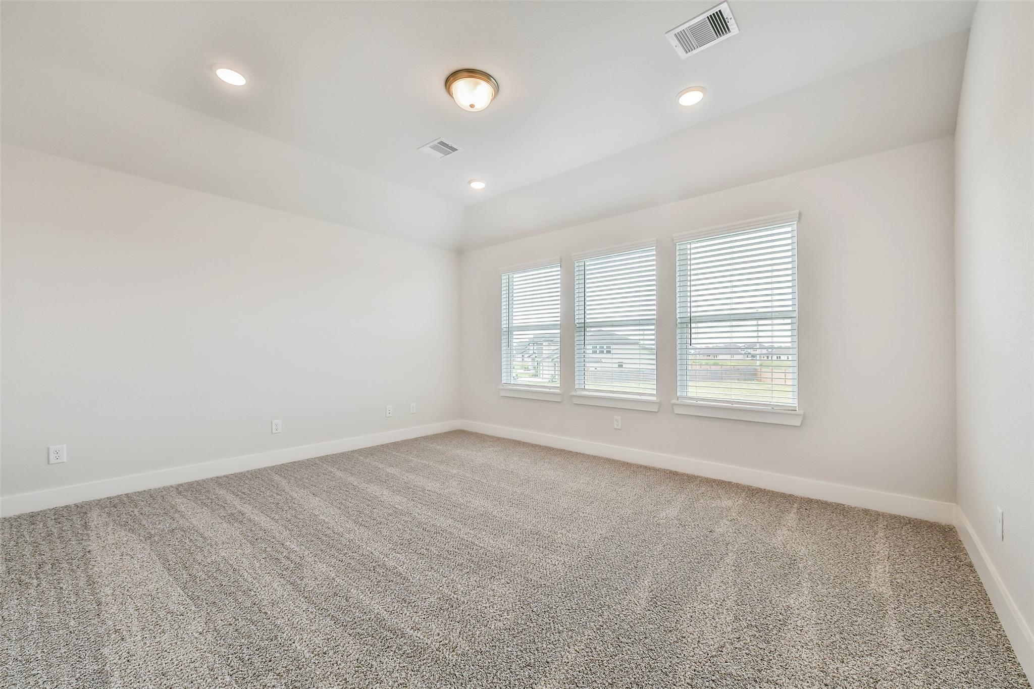 Spacious empty bedroom with white walls, beige carpet, large windows with blinds, and recessed lighting in Davidson Homes The Victoria A, Lago Mar, Texas City