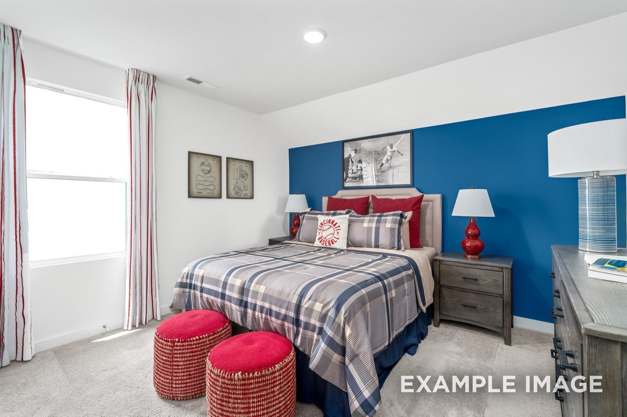 Cozy bedroom with blue accent wall, gray plaid bedding, red pillows, ottomans, and curtains in Davidson Homes The Ash A, Gallatin, TN
