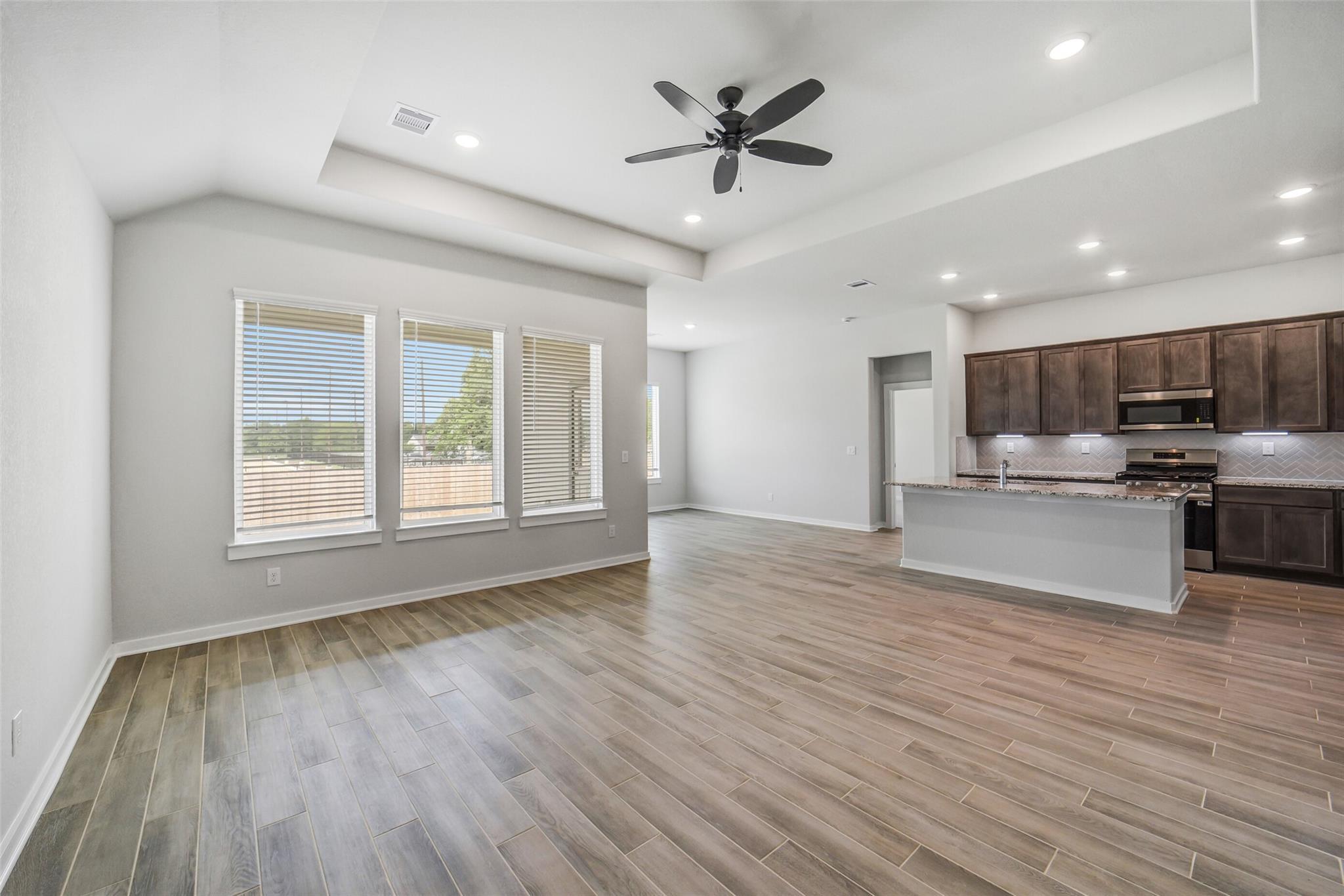 Open-concept kitchen and living area with brown shaker cabinets, stainless appliances, island, ceiling fan, and large windows in Davidson Homes Acadia C, Magnolia TX