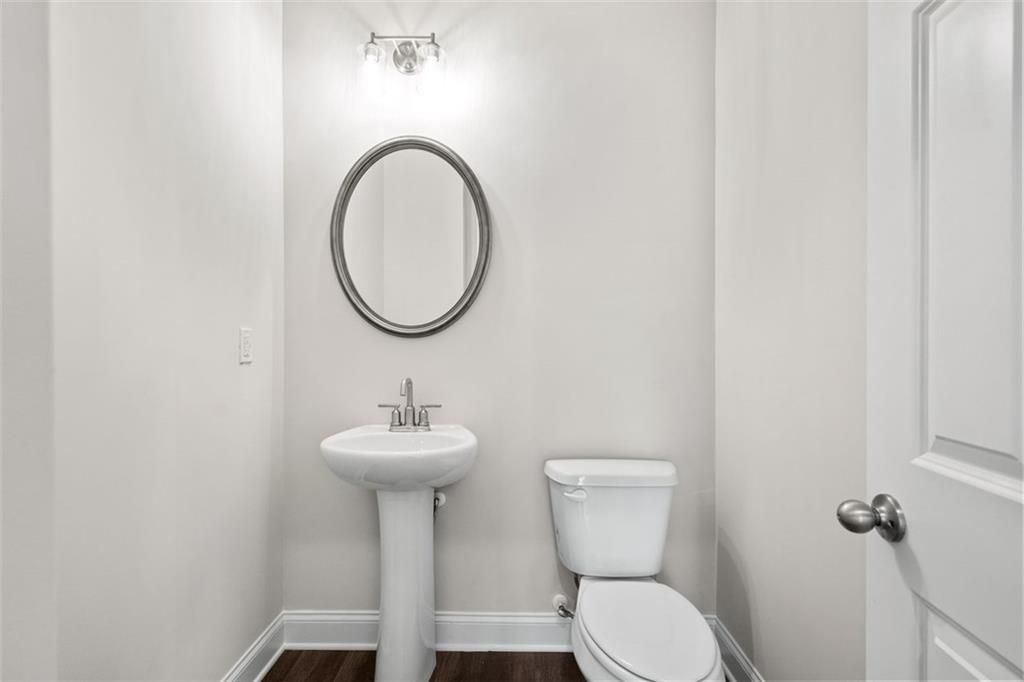 Elegant powder room featuring pedestal sink, round mirror, and toilet in Davidson Homes The Ash C at Wehunt Meadows, Hoschton, Georgia