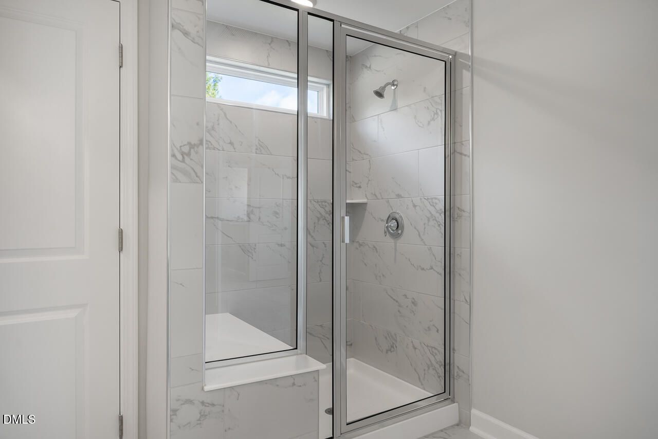 Elegant frameless glass shower with white marble tiles, built-in bench, and window in master bath of Davidson Homes The Graham in Fuquay-Varina, NC
