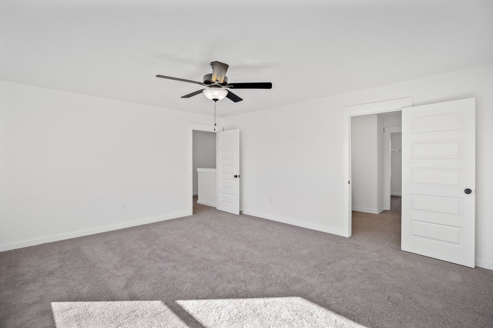 Spacious empty bedroom with white walls, beige carpet, ceiling fan, and open doors to closet and stairs in Davidson Homes The Willow, Gallatin, Tennessee