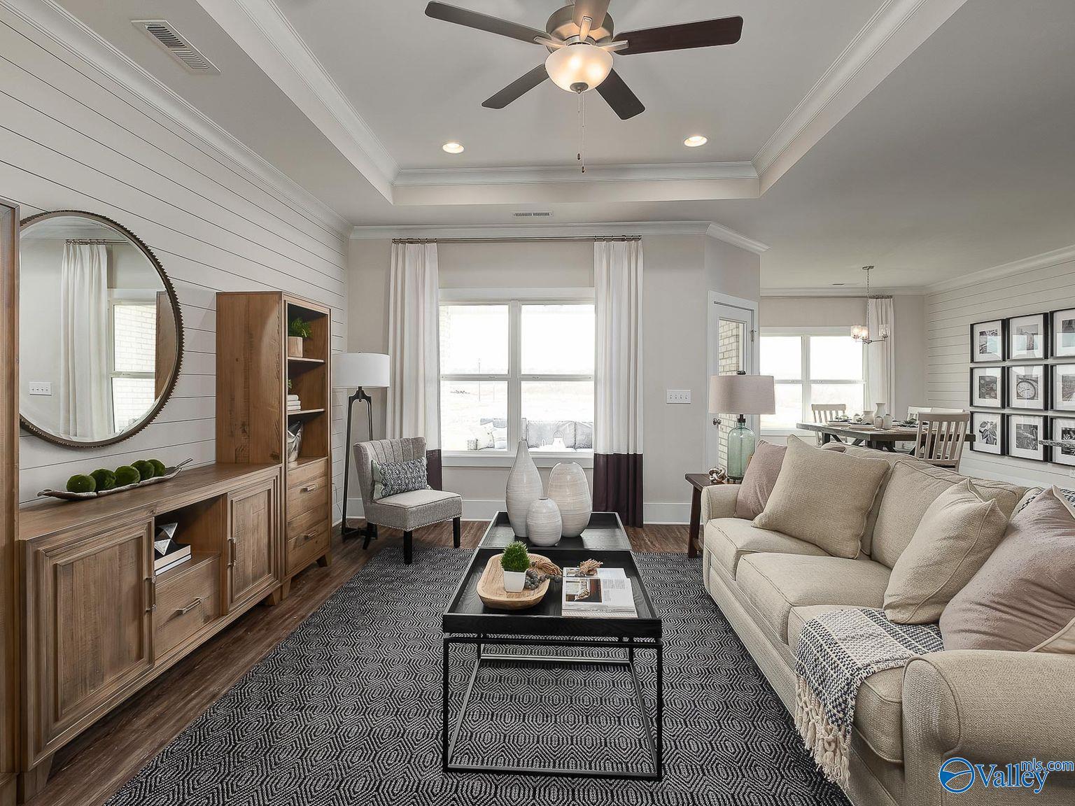 Cozy living room with beige sofa, wooden cabinet, large windows, and ceiling fan in Davidson Homes The Everett, Toney, Alabama