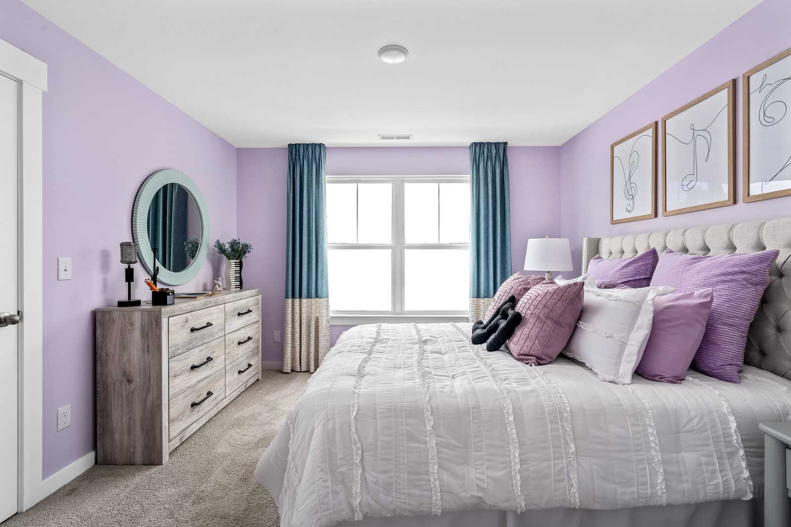 Lavender bedroom at Woods Crossing in Gallatin TN with tufted bed, purple pillows, wooden dresser, round mirror, and blue curtains