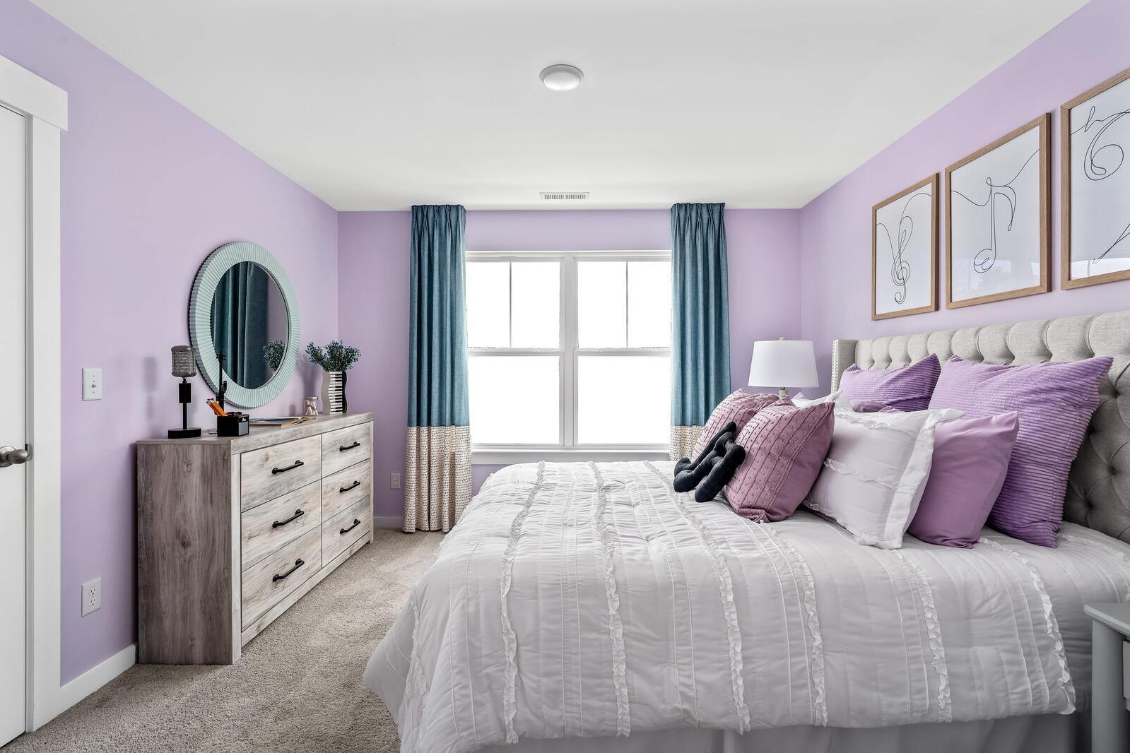 Lavender bedroom at Woods Crossing in Gallatin TN with tufted bed, purple pillows, wooden dresser, round mirror, and blue curtains