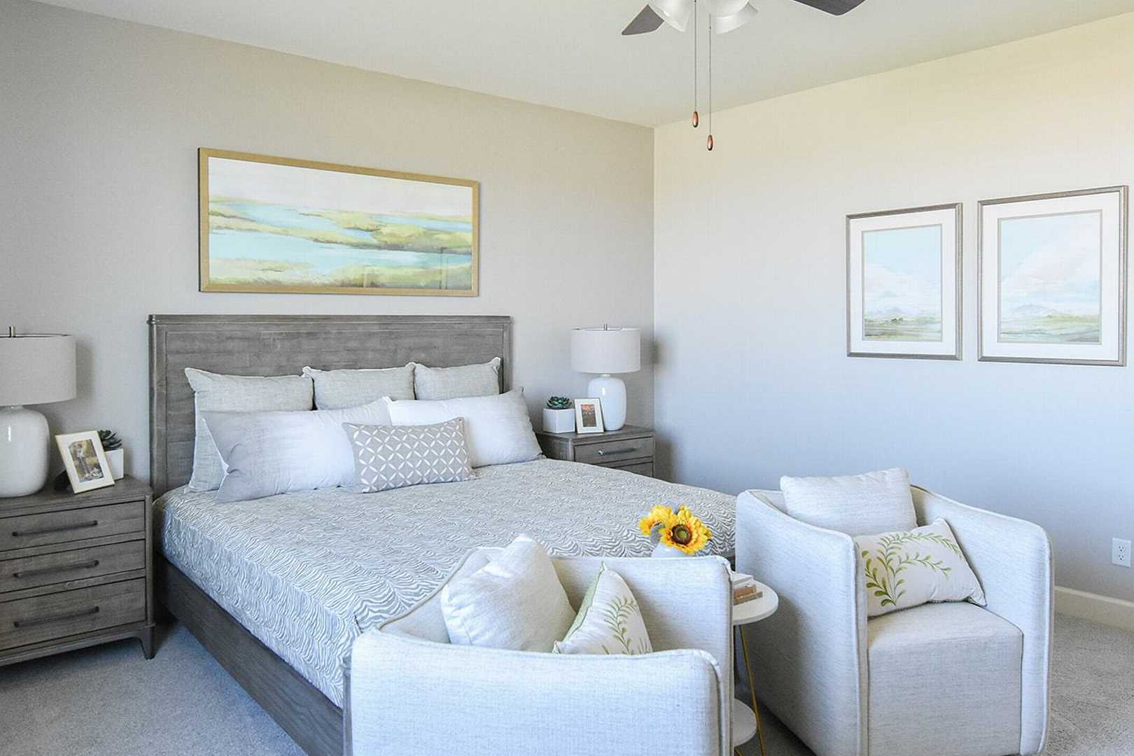 Spacious master bedroom in The Summit B featuring king bed with gray headboard, sunflowers, armchairs, and abstract art on beige walls