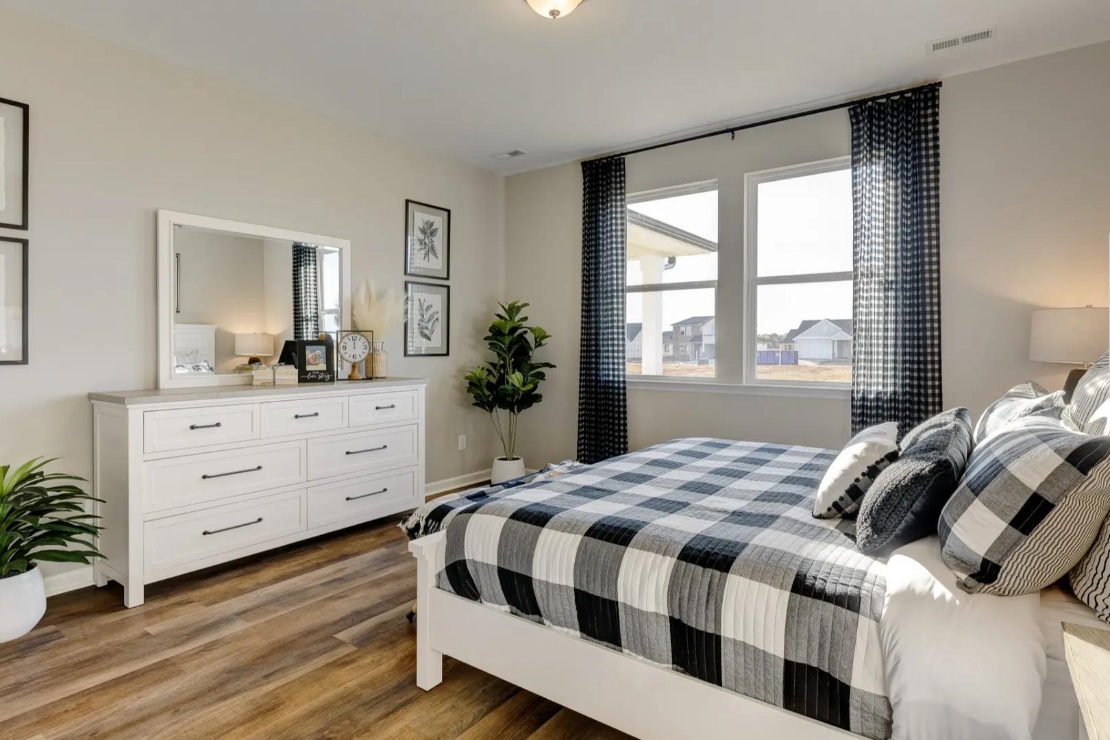 Spacious master bedroom in The Aurora A with checkered black-and-white bedding, white dresser, potted plants, and botanical wall art