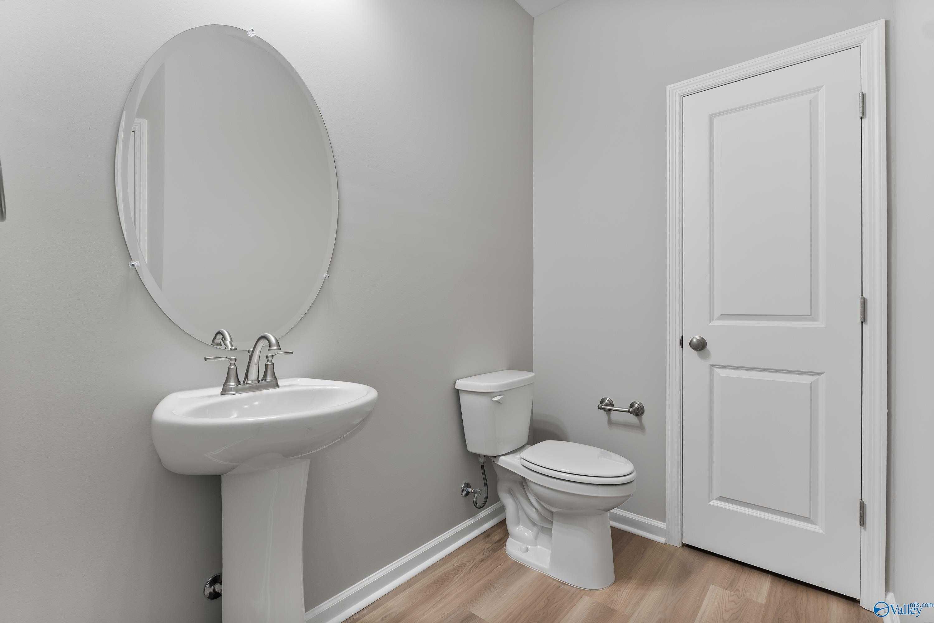 Modern powder room featuring white pedestal sink, oval mirror, toilet, and gray walls in Evermore Homes The Grace, Madison, Alabama
