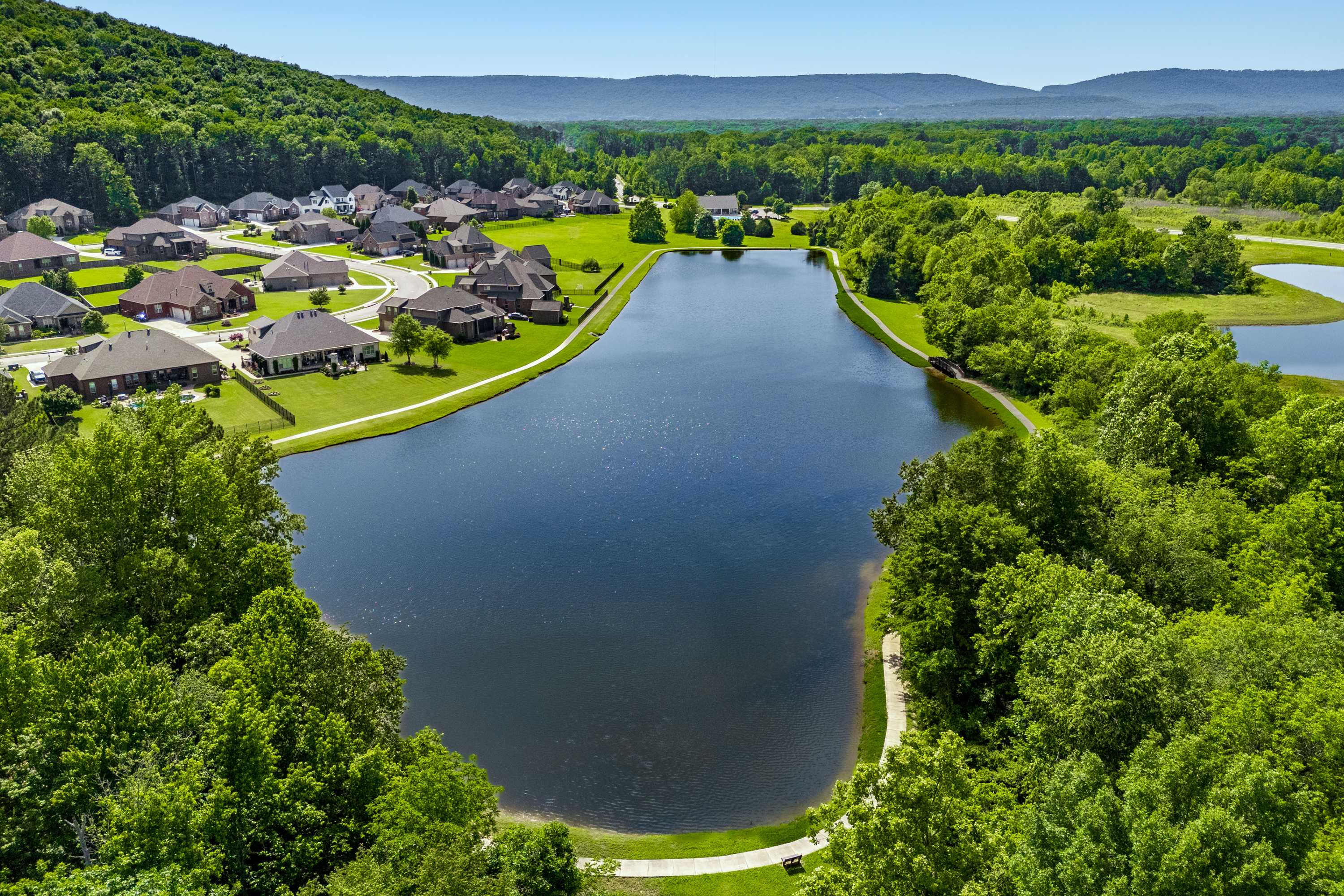Aerial view of serene pond with walking path, modern homes, and lush greenery at The Meadows at Hampton Cove in Owens Cross Roads, Alabama