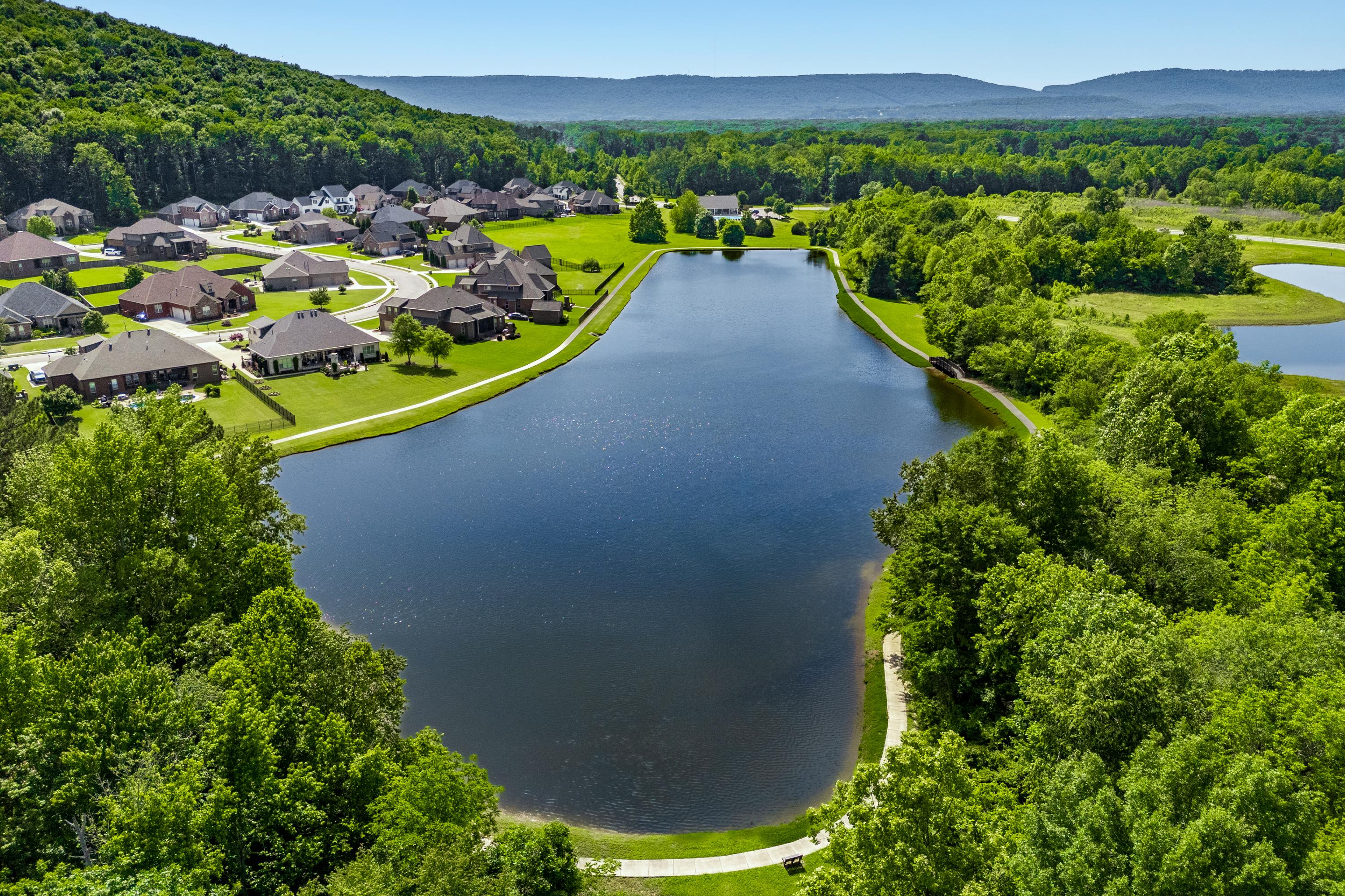 Aerial view of serene pond with walking path, modern homes, and lush greenery at The Meadows at Hampton Cove in Owens Cross Roads, Alabama