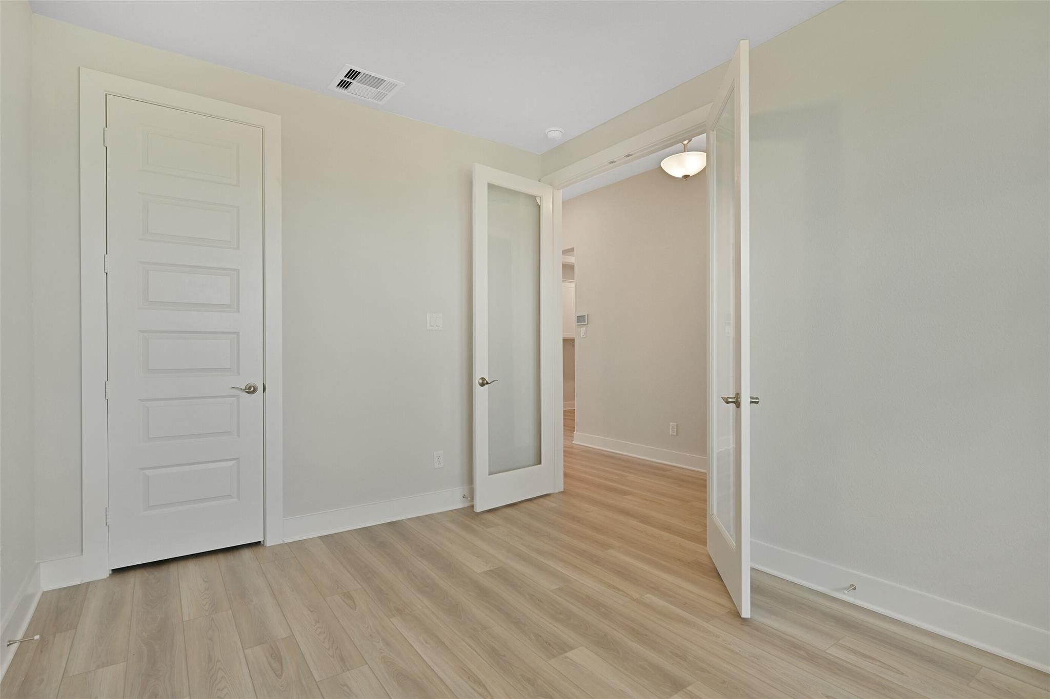 Spacious empty bedroom with light oak floors, open glass-paneled door to bathroom, neutral walls in Davidson Homes The Edward A, Lago Mar, Texas
