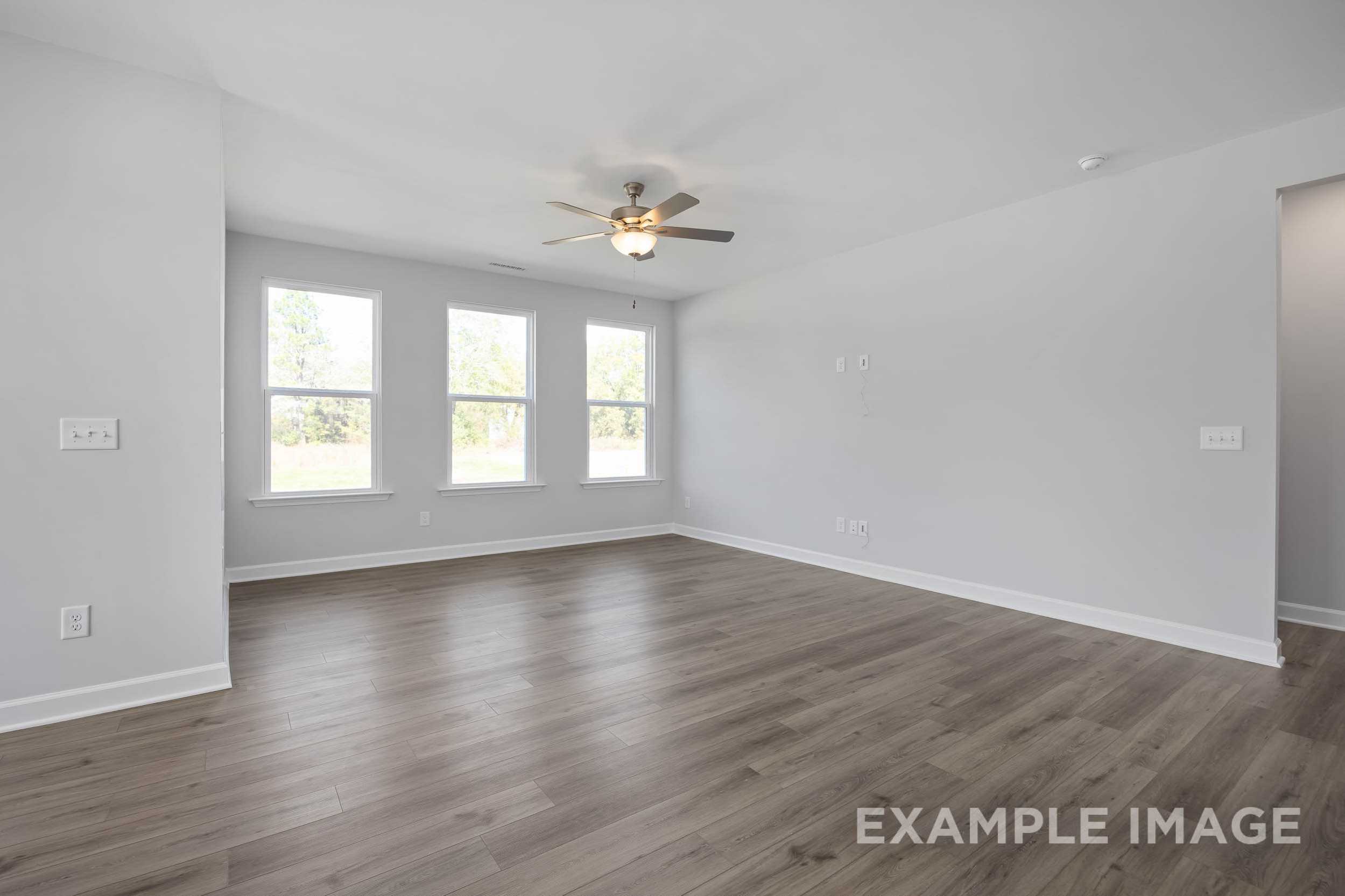 Spacious empty living room in The Ash home design with large triple windows, ceiling fan, light gray walls, and hardwood floors