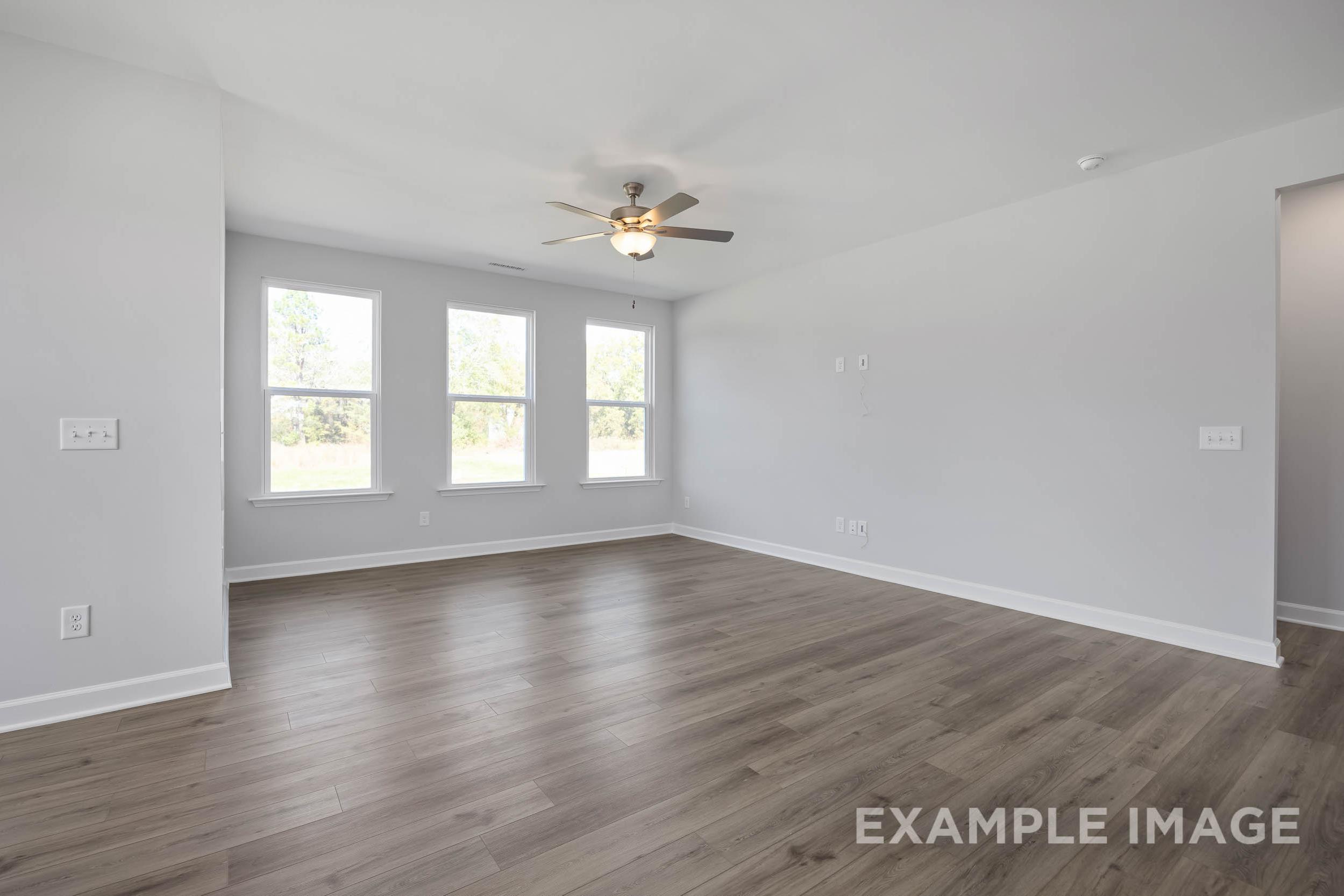 Spacious empty living room in The Ash home design with large triple windows, ceiling fan, light gray walls, and hardwood floors