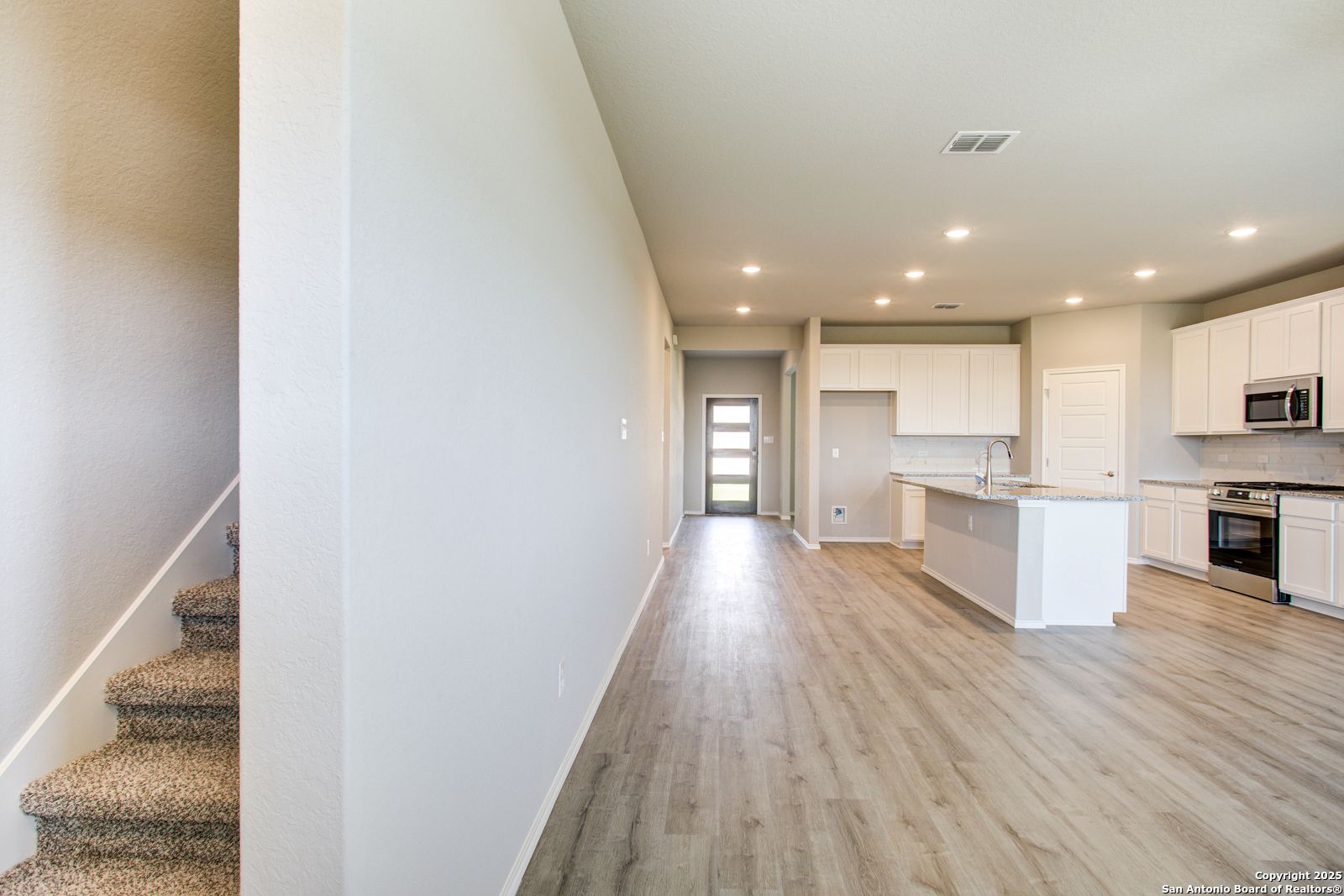 Spacious hallway with carpeted staircase opening to modern kitchen with white cabinets and island in Davidson Homes The Douglas G, San Antonio