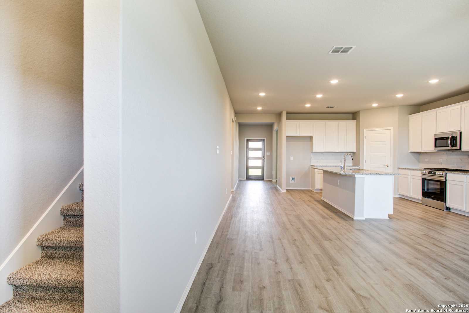 Spacious hallway with carpeted staircase opening to modern kitchen with white cabinets and island in Davidson Homes The Douglas G, San Antonio