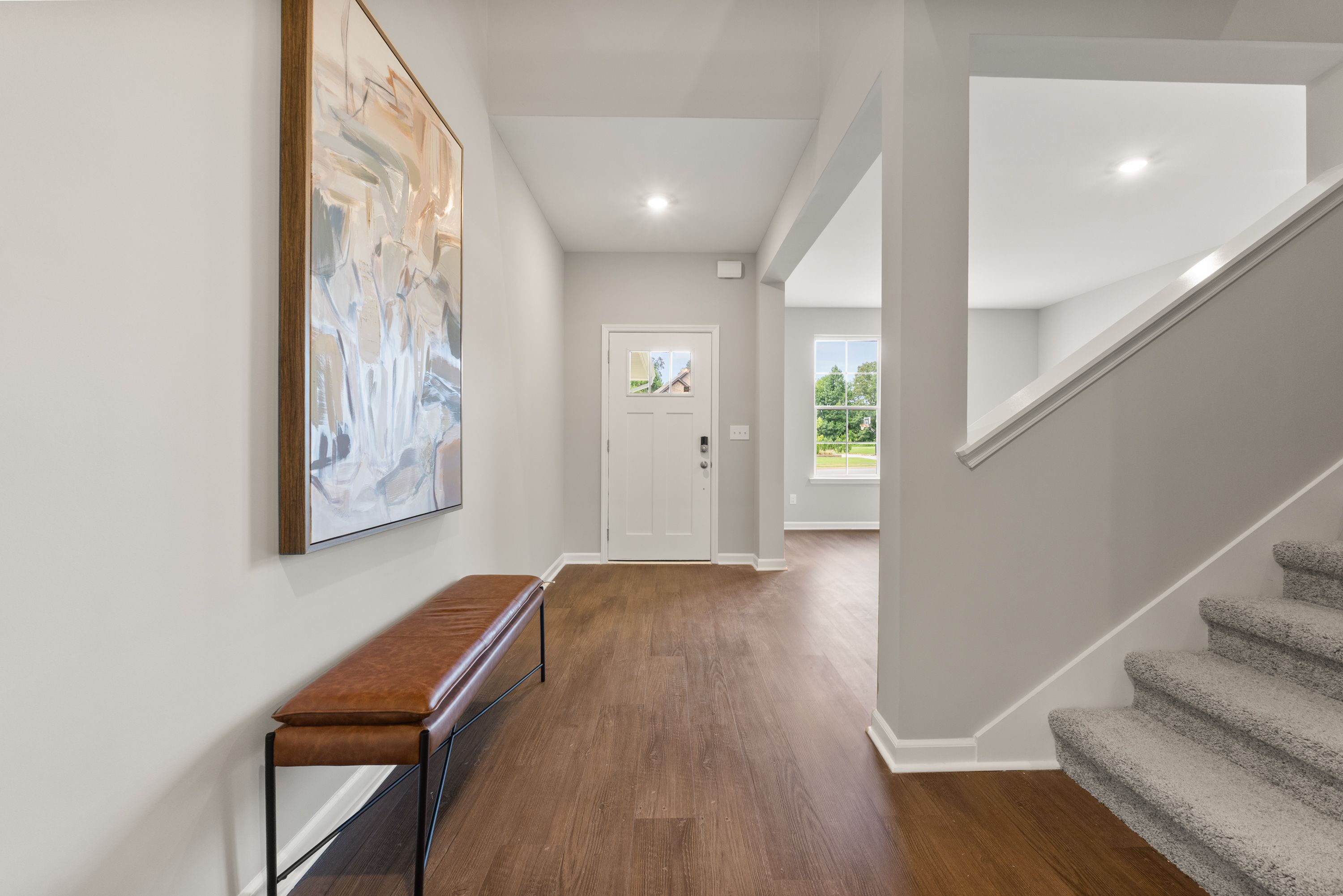Bright open hallway in The Avalon E home by Evermore Homes with abstract wall art, leather bench, white door, and carpeted staircase
