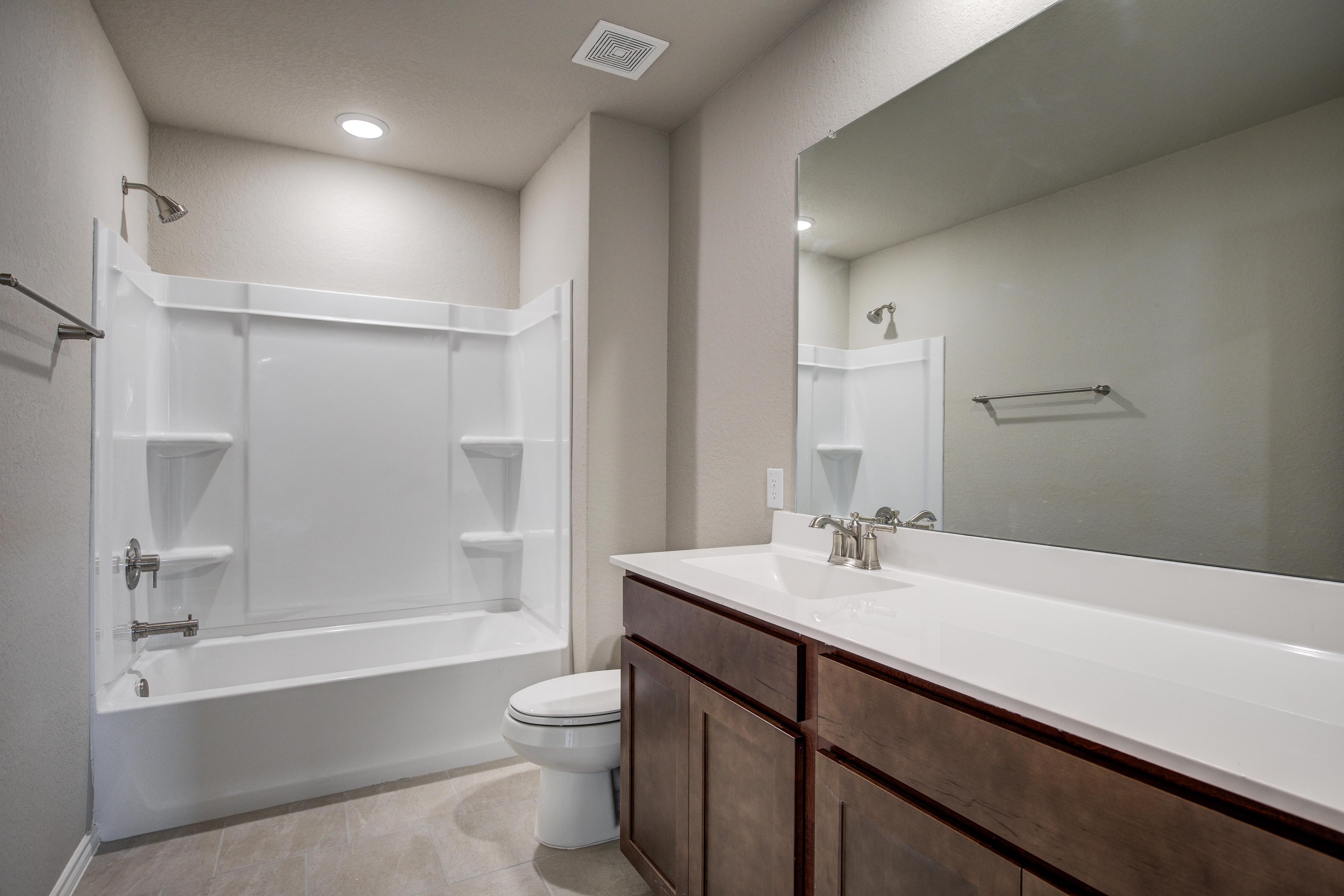 Spacious bathroom in The Murray G featuring white tub-shower combo, brown shaker vanity, large mirror, Davidson Homes San Antonio