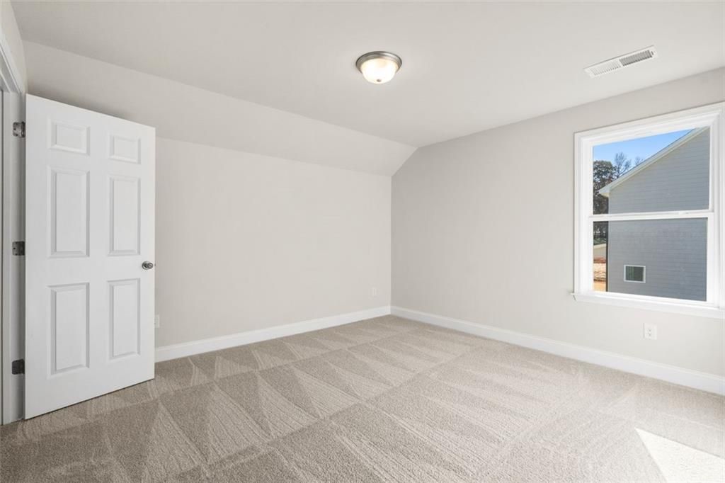 Bright secondary bedroom with sloped ceiling, light gray walls, white door, and patterned carpet in The Ash C at Wehunt Meadows, Hoschton