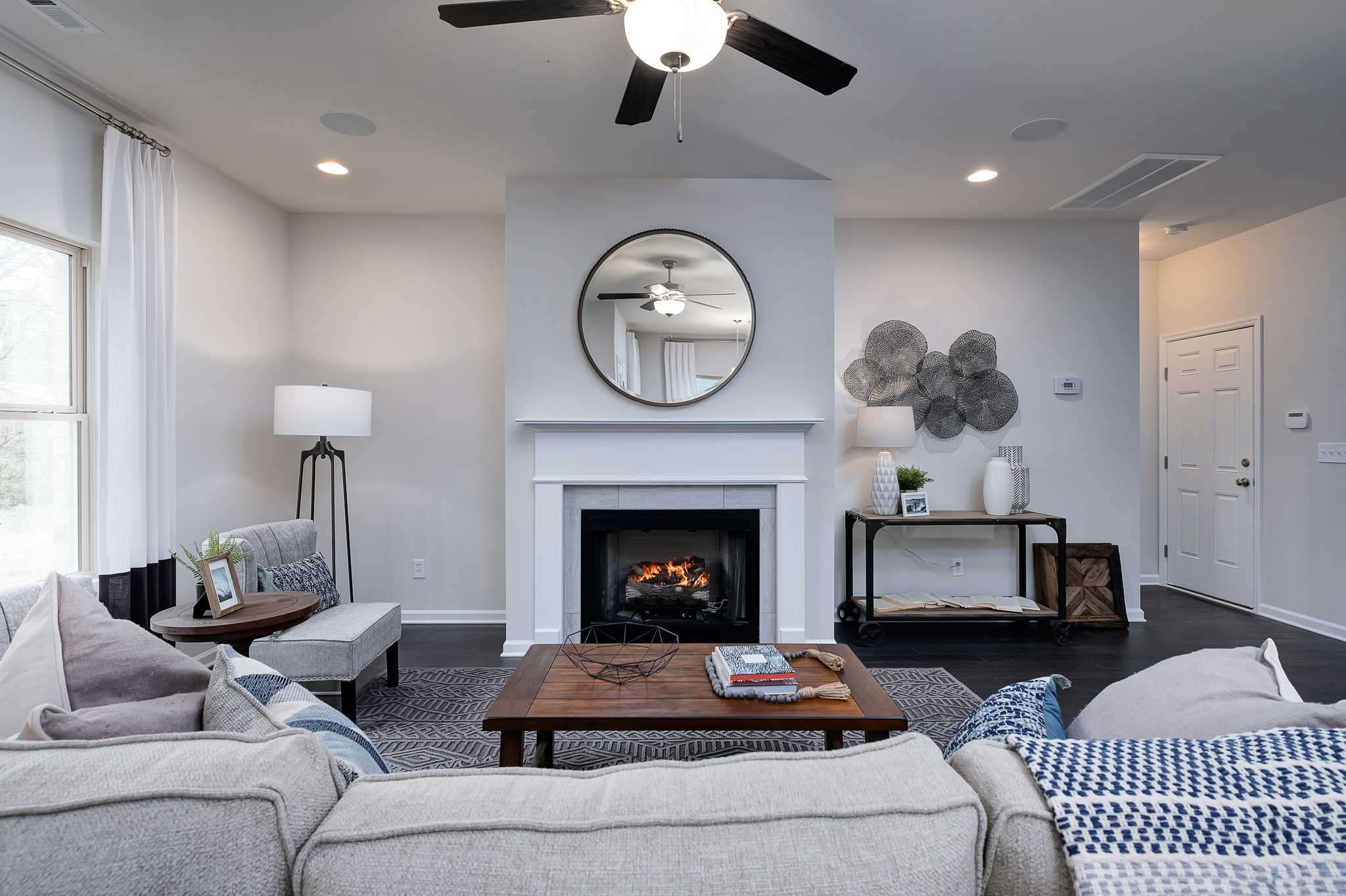 Modern living room with white fireplace, round mirror, gray sofa, coffee table, and ceiling fan at The Reserve at Overton in Hampton Cove AL