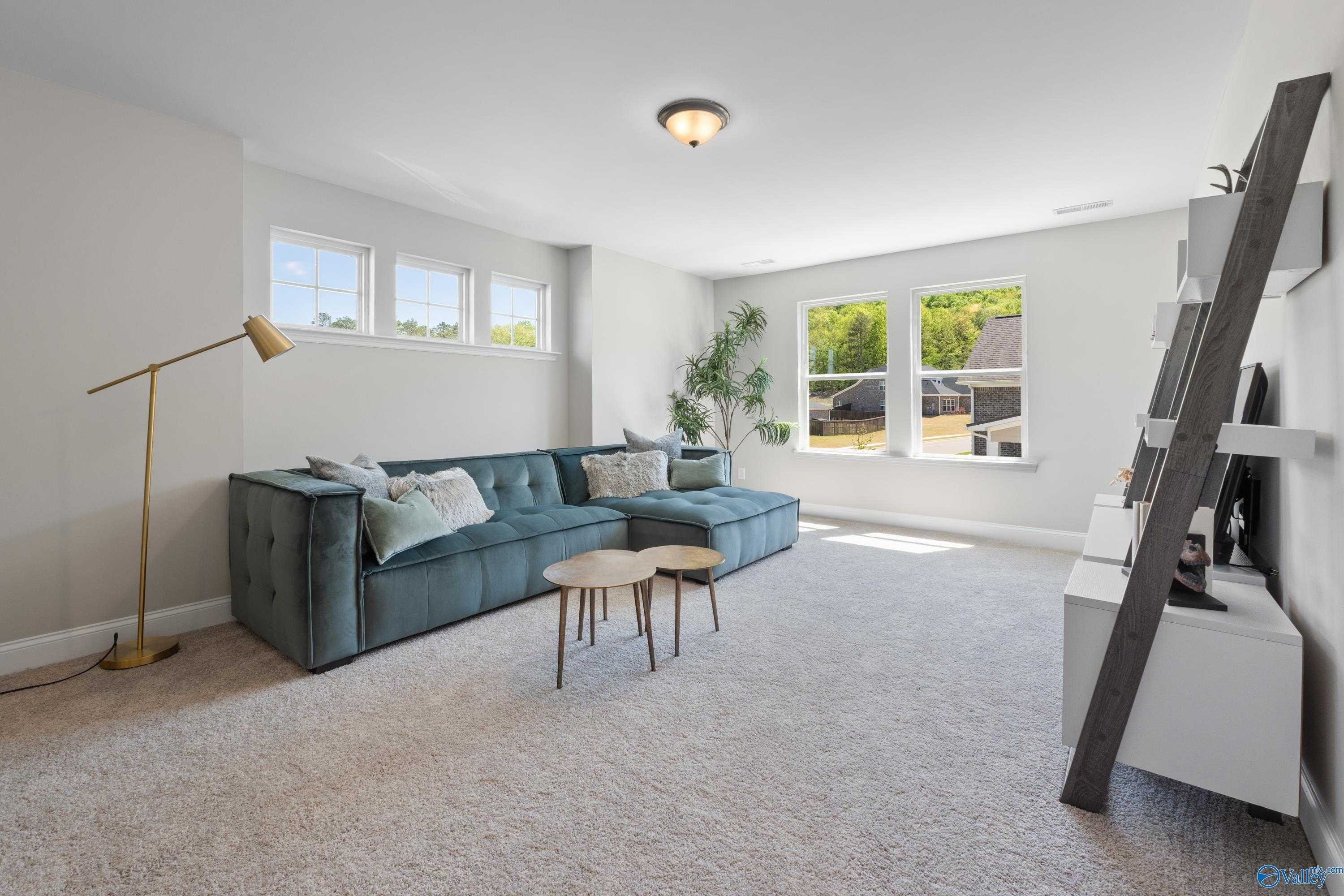 Cozy living room with blue velvet sectional sofa, ladder shelf, potted plants, and large windows in 5-bedroom Evermore Homes The Haven, Owens Cross Roads, Alabama
