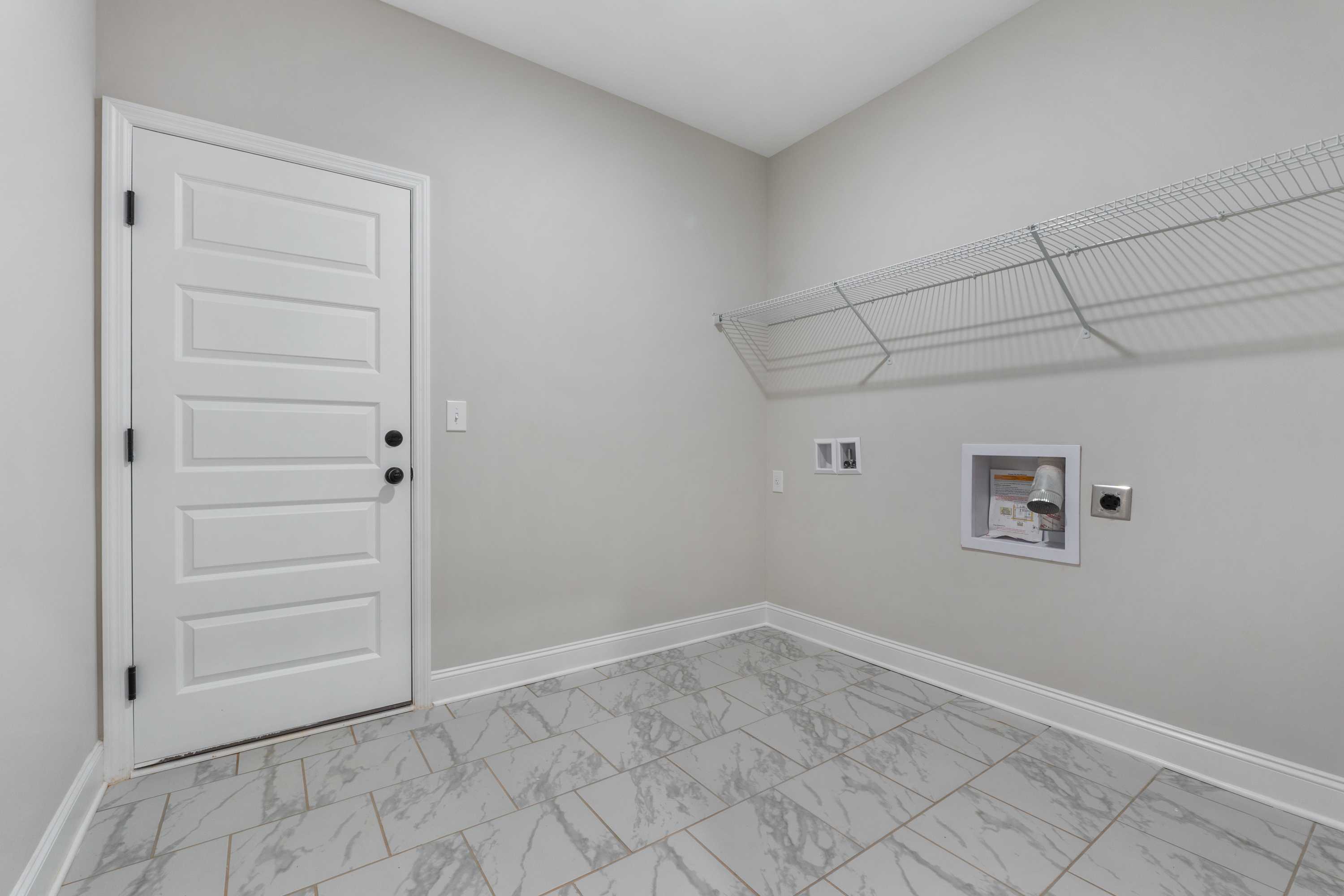 Spacious laundry room in The Oxford home design with white door, wire shelving, washer dryer hookups, and gray tile floor