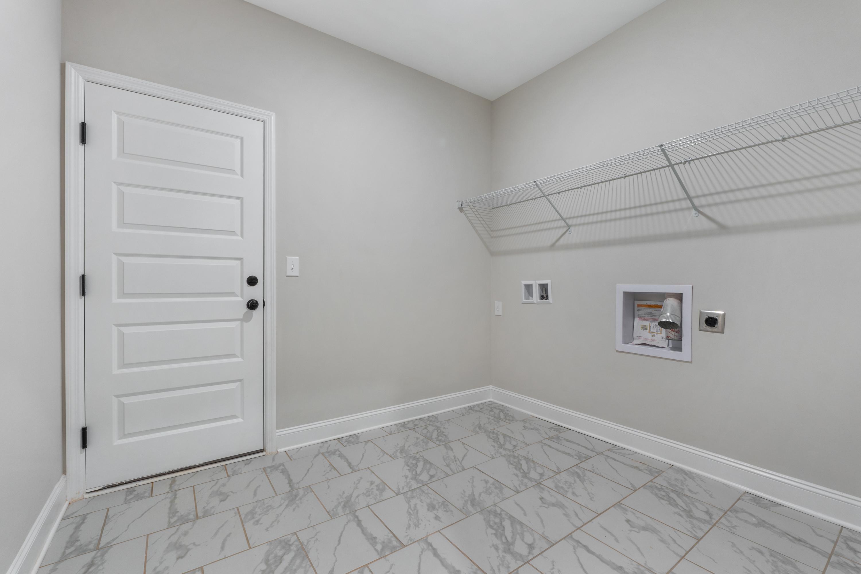 Spacious laundry room in The Oxford home design with white door, wire shelving, washer dryer hookups, and gray tile floor