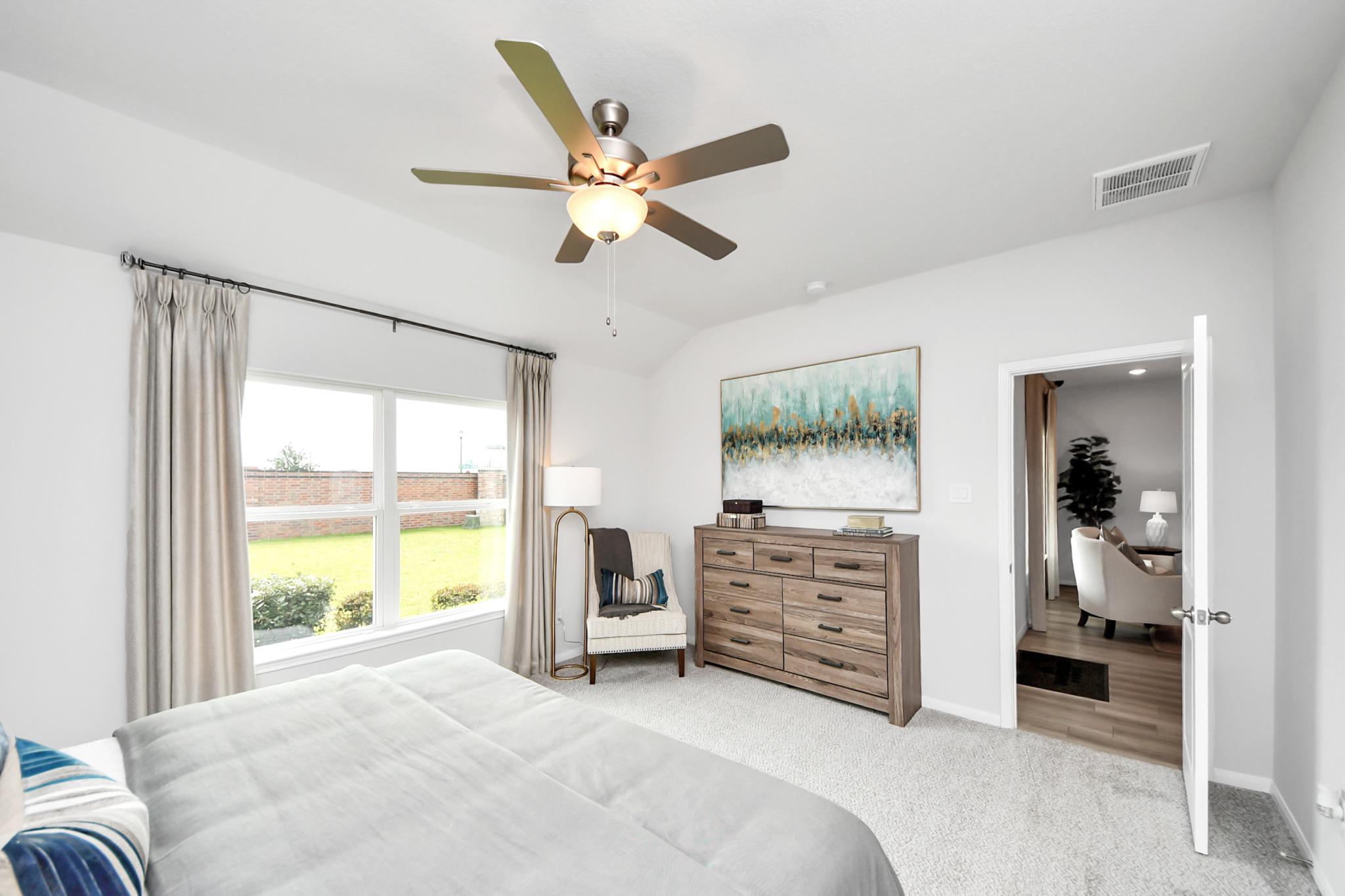 Bright bedroom in Sunterra, Katy, Texas by Davidson Homes featuring a ceiling fan, wood dresser, and view of the backyard.