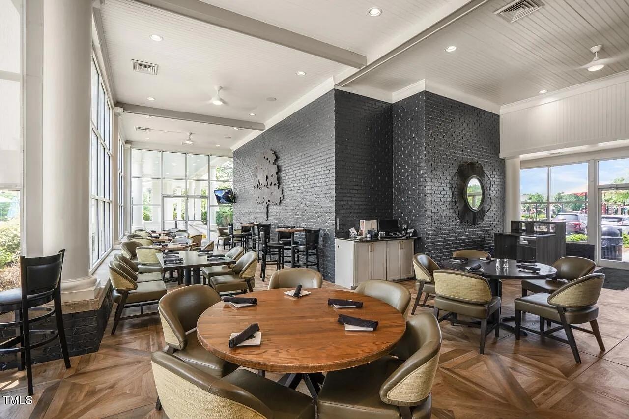 Spacious clubhouse lounge at Addison West in Holly Springs NC with round wooden tables, upholstered chairs, brick accent wall, and large windows