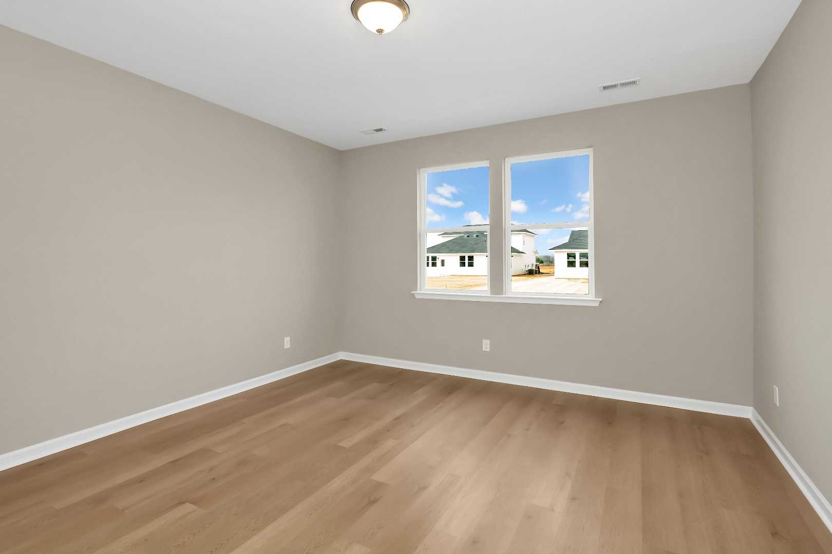 Spacious bedroom in The Orion home with light gray walls, oak hardwood floors, and sunny window views of neighborhood homes