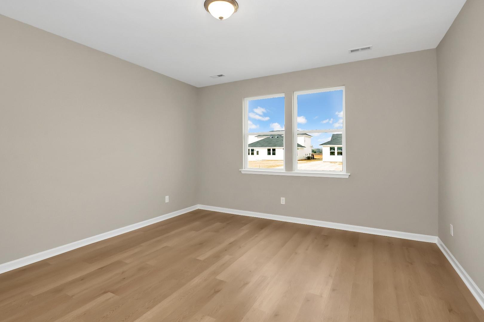 Spacious bedroom in The Orion home with light gray walls, oak hardwood floors, and sunny window views of neighborhood homes