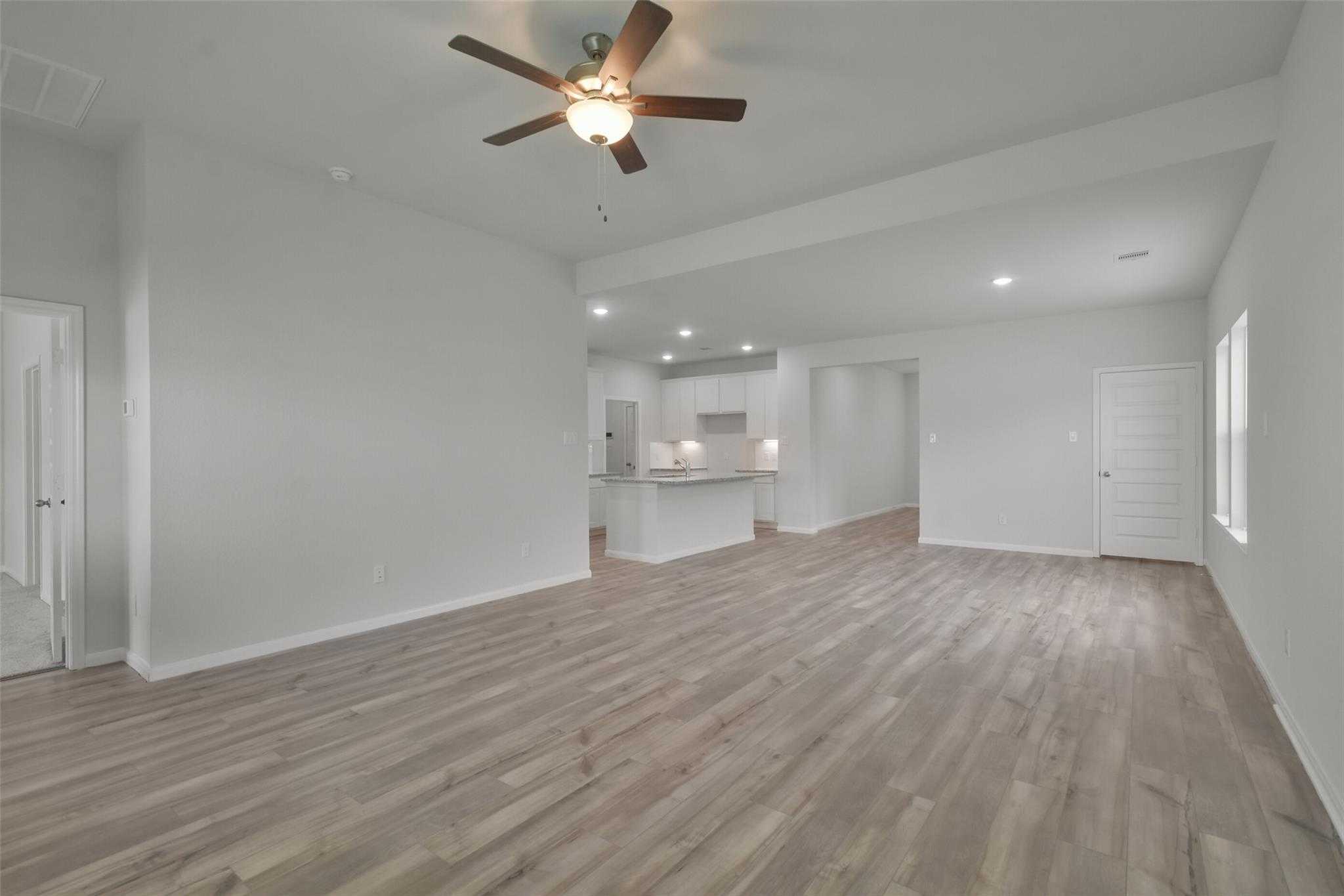 Spacious open living room with hardwood floors, ceiling fan, and adjacent white kitchen in Davidson Homes The Tierra A, Dayton, Texas