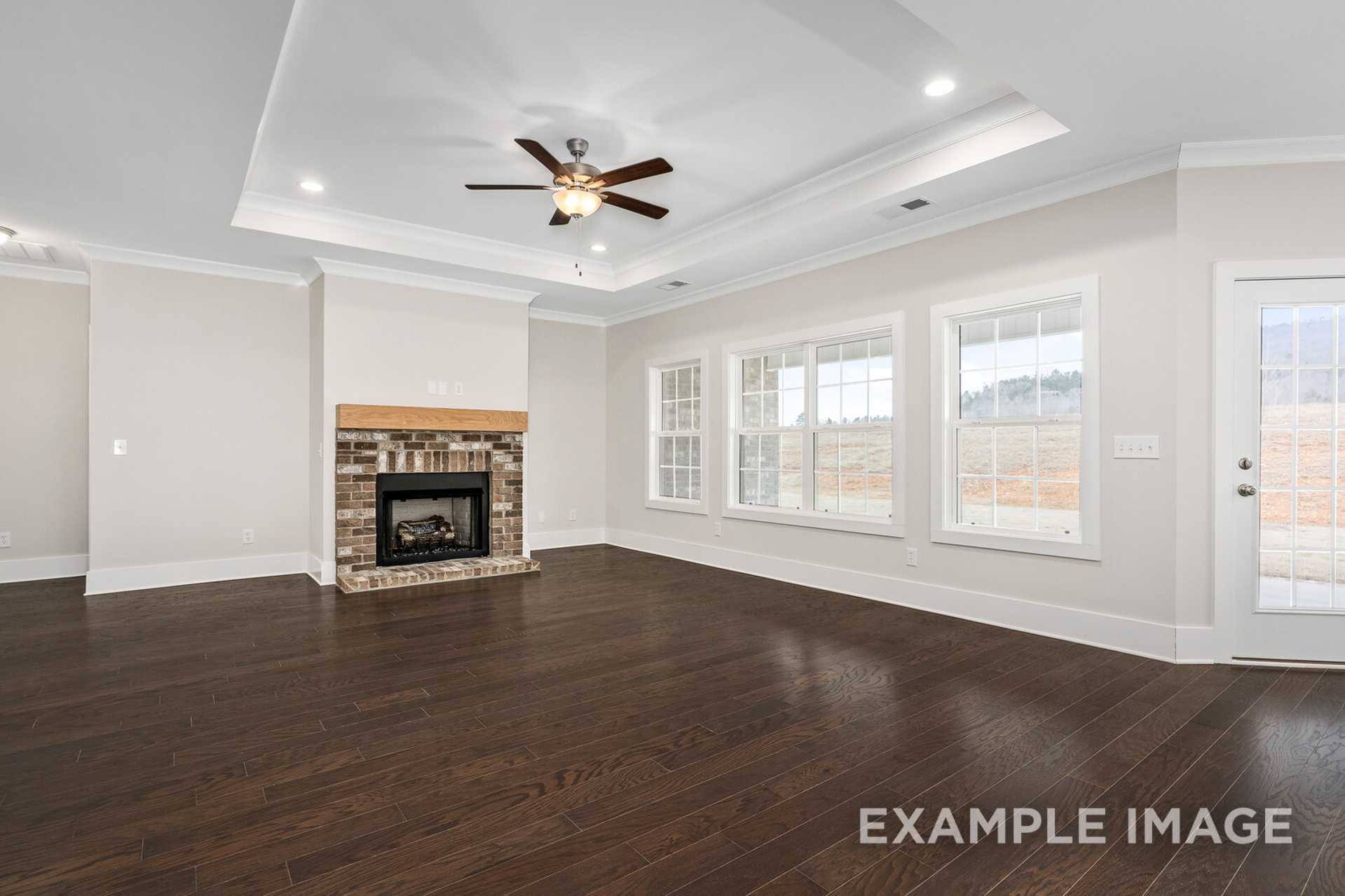 Spacious living room in The Rockford home design featuring brick fireplace, hardwood floors, ceiling fan, and large windows