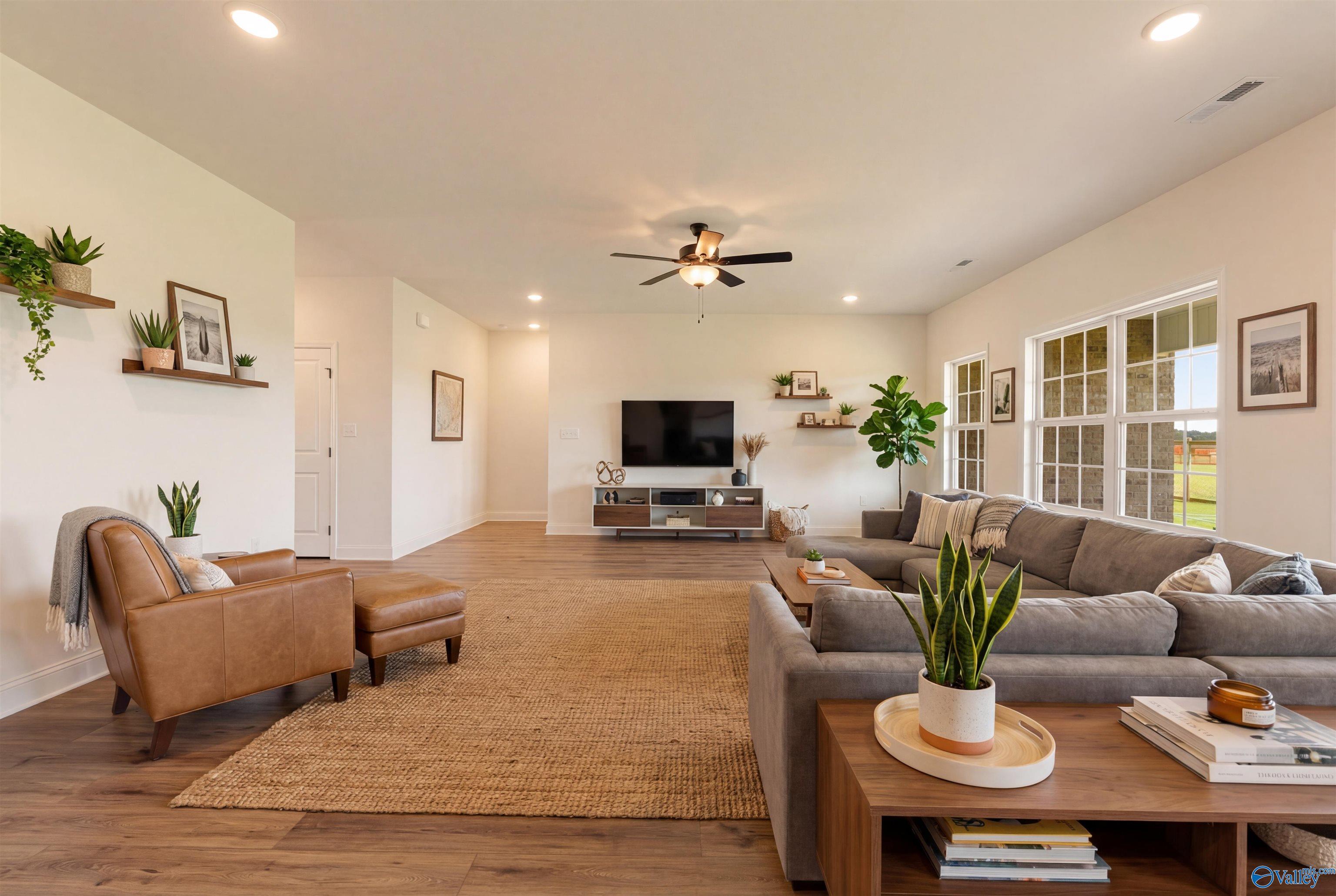 Spacious living room with gray L-shaped sofa, leather armchair, TV console, plants, and large windows in The Rockford B, Toney, Alabama