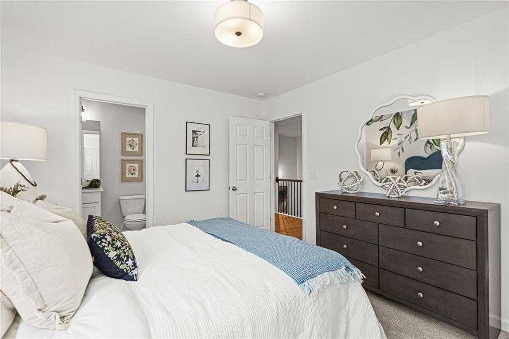 Cozy bedroom with plush white bed, blue throw, wooden dresser, and open bathroom door in Davidson Homes The Hickory B, Hoschton, GA