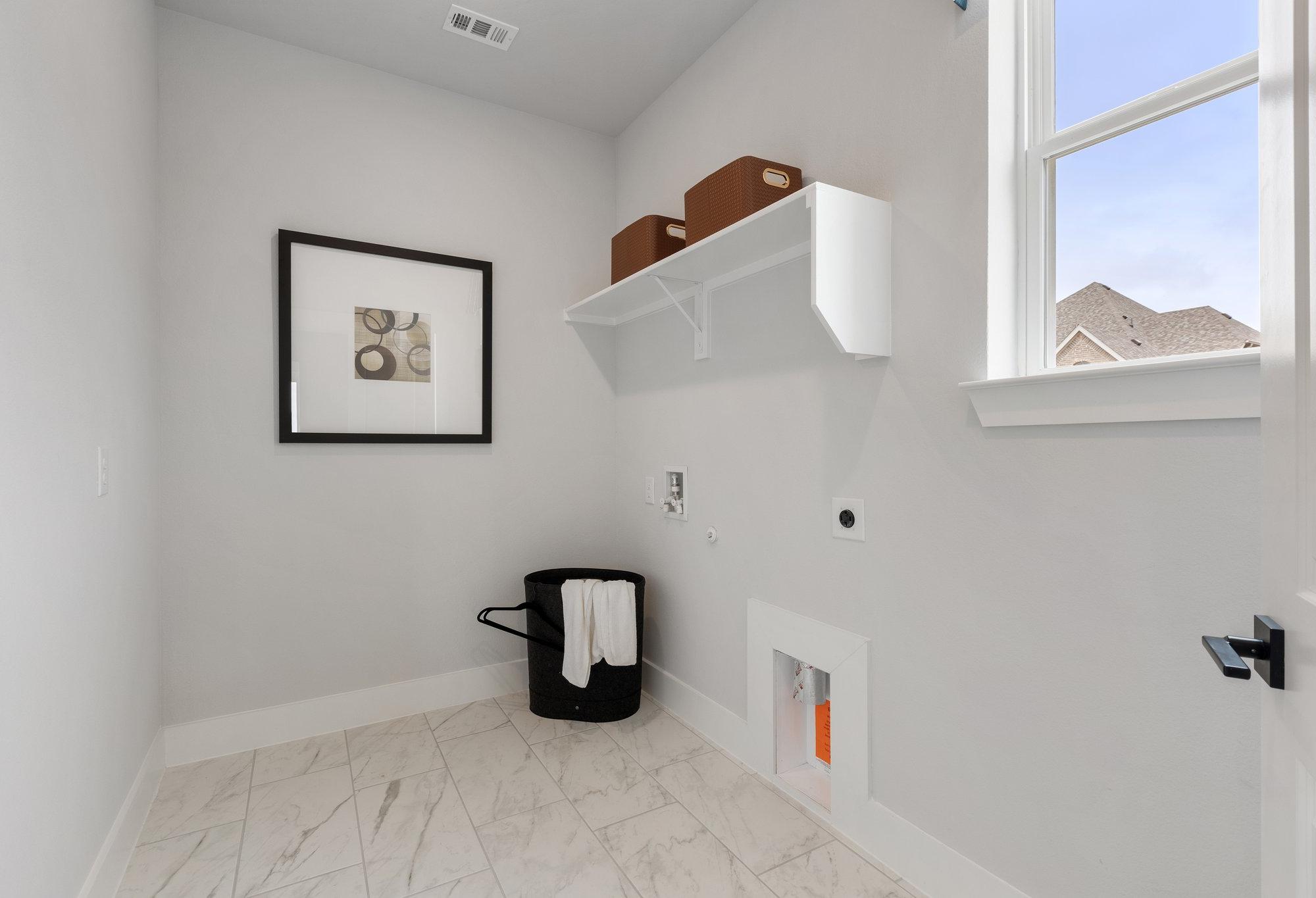 Spacious laundry room in The Sequoia MA home with white shelves, brown storage bins, framed abstract art, black basket, washer-dryer hookups, and window view