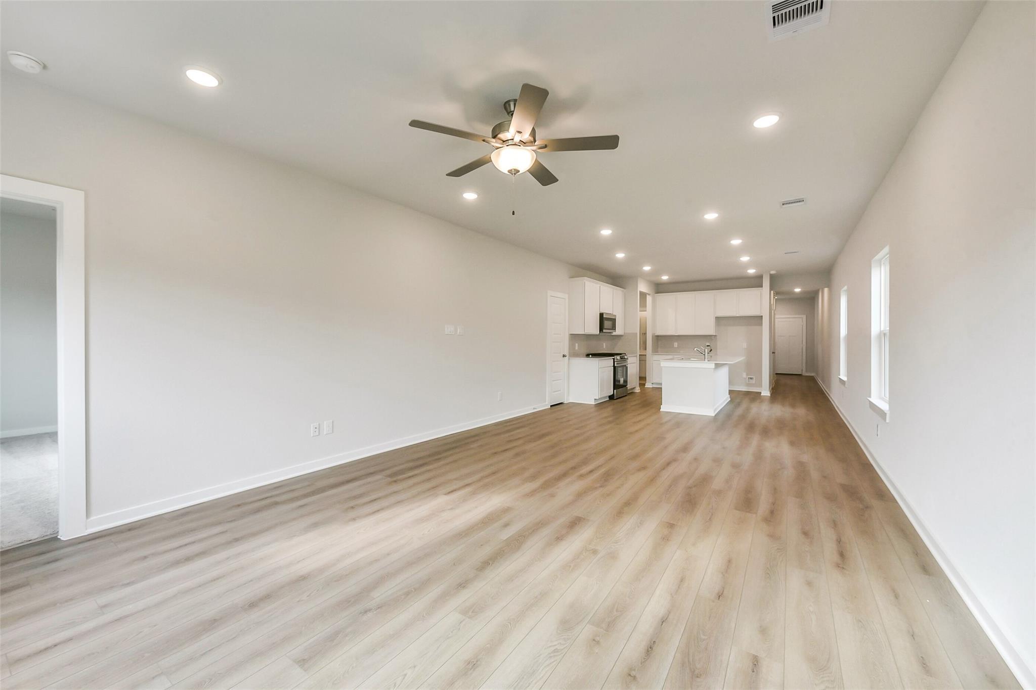 Open hallway with laminate flooring, white walls, ceiling fan and recessed lights leading to modern white kitchen in The Frio G home, Dayton Texas