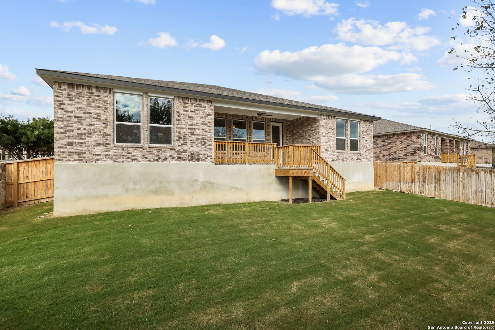 Backyard view of The Lanier H single-story brick home with covered porch, wooden deck stairs, and lush green lawn in Ladera, San Antonio by Davidson Homes