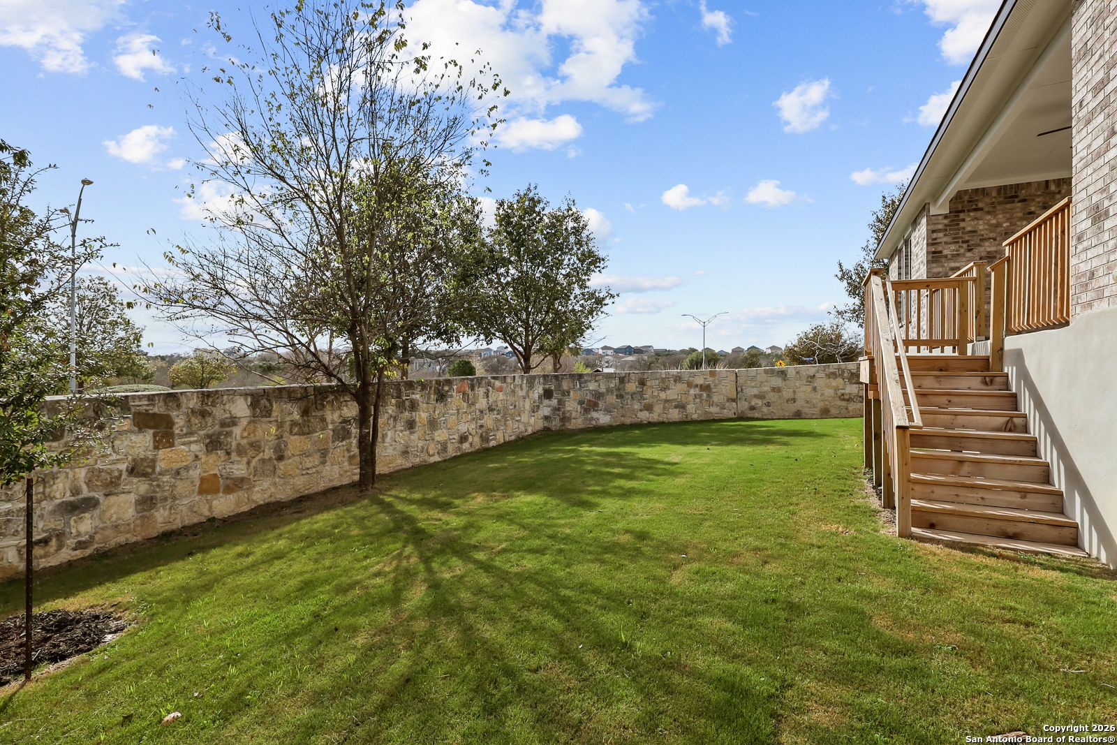 Private backyard with lush green lawn, curved stone wall, wooden stairs, and trees in Davidson Homes The Lanier H, Ladera, San Antonio