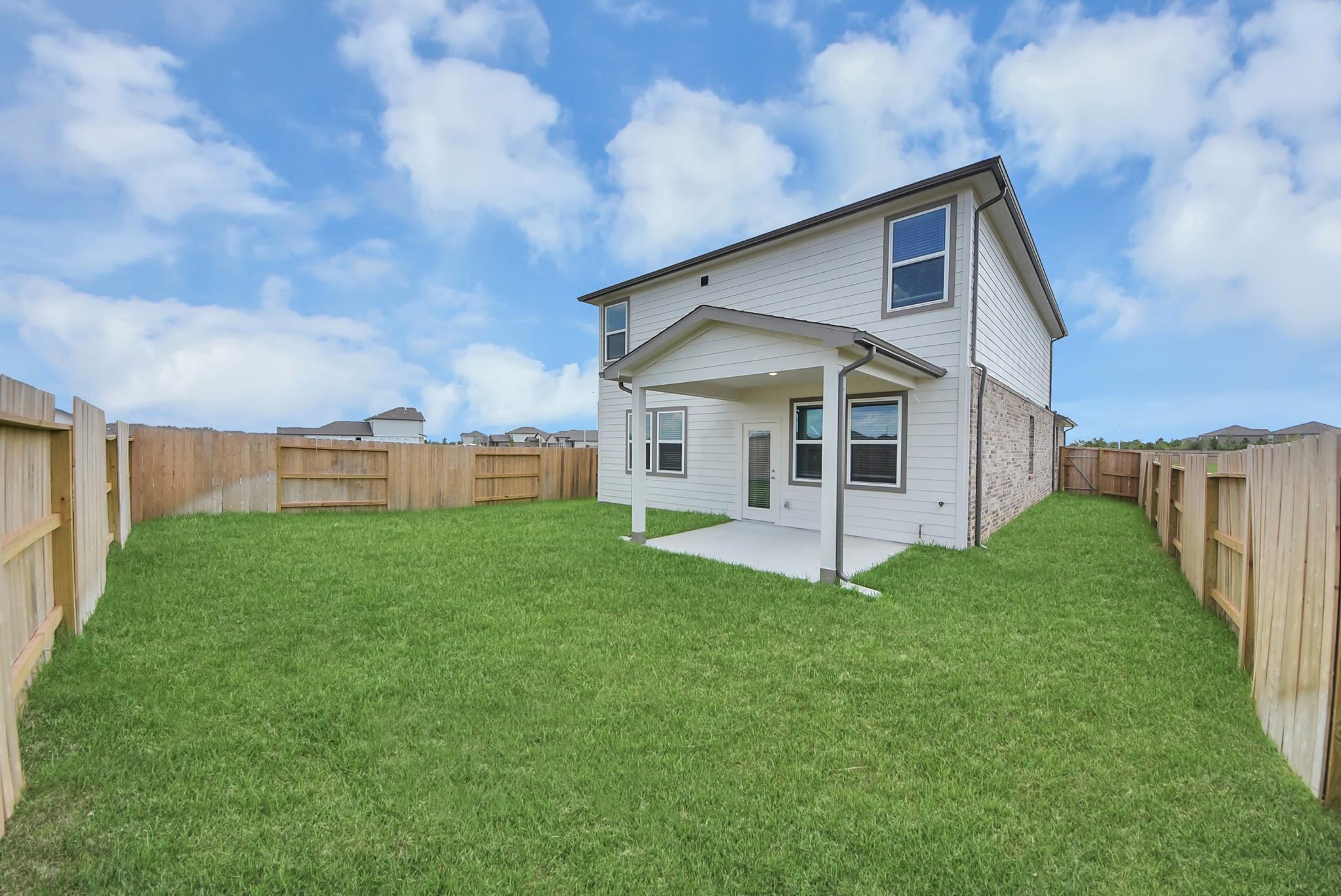 Two-story home exterior with covered patio, fenced backyard, and lush green lawn at Davidson Homes The San Marcos E in Beasley, Texas