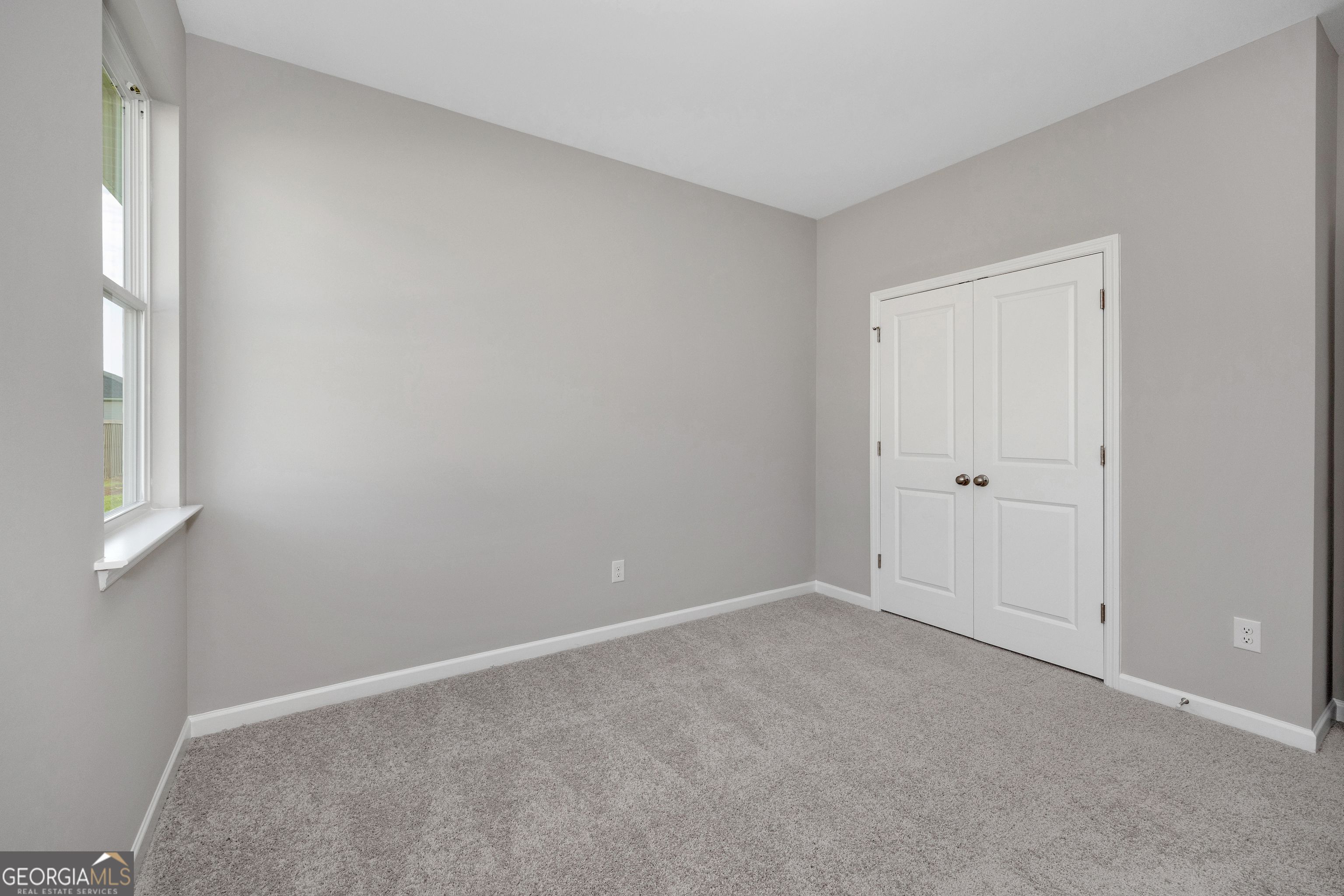 Empty bedroom with gray walls, plush carpet, double white closet doors, and large window in Evermore Homes The Luna, Ivy Glen, Perry, Georgia